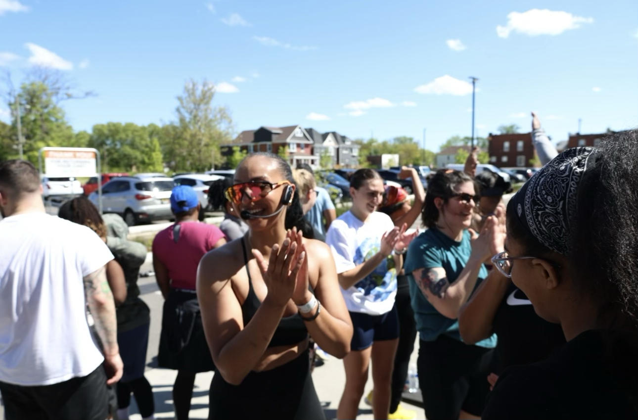 Group of people outdoors on a sunny day, clapping and celebrating, with parking lot and buildings in the background.