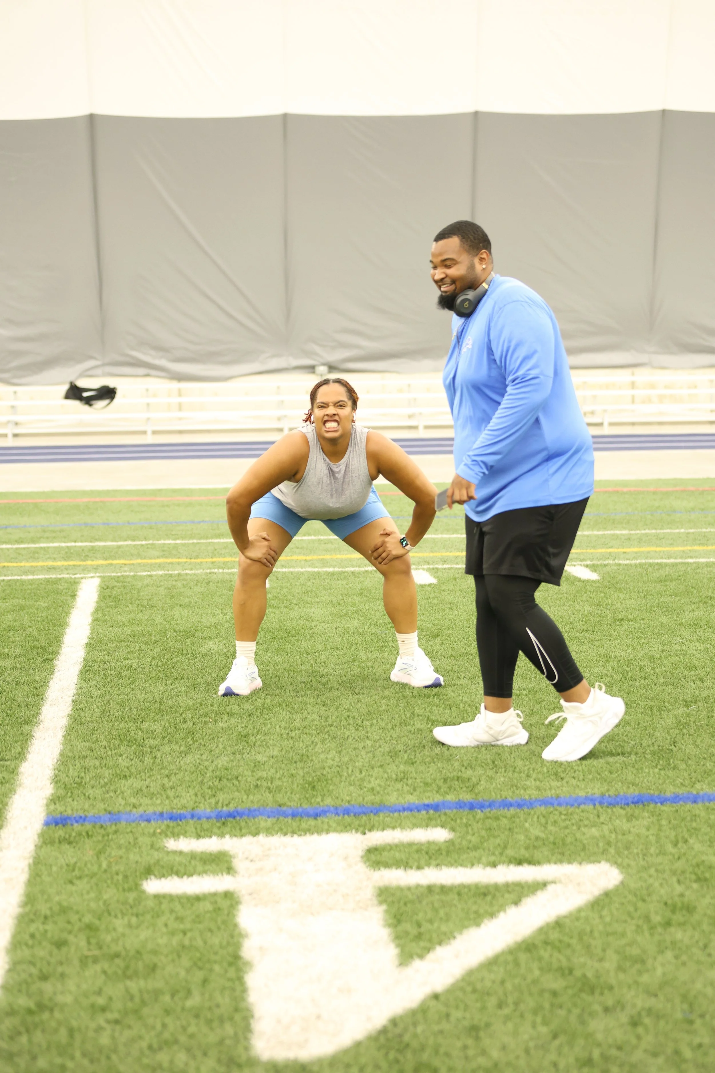 A woman in athletic clothing squatting with a grimace on her face while a man in athletic clothing with headphones standing nearby appears to be coaching or encouraging her on an indoor sports field.
