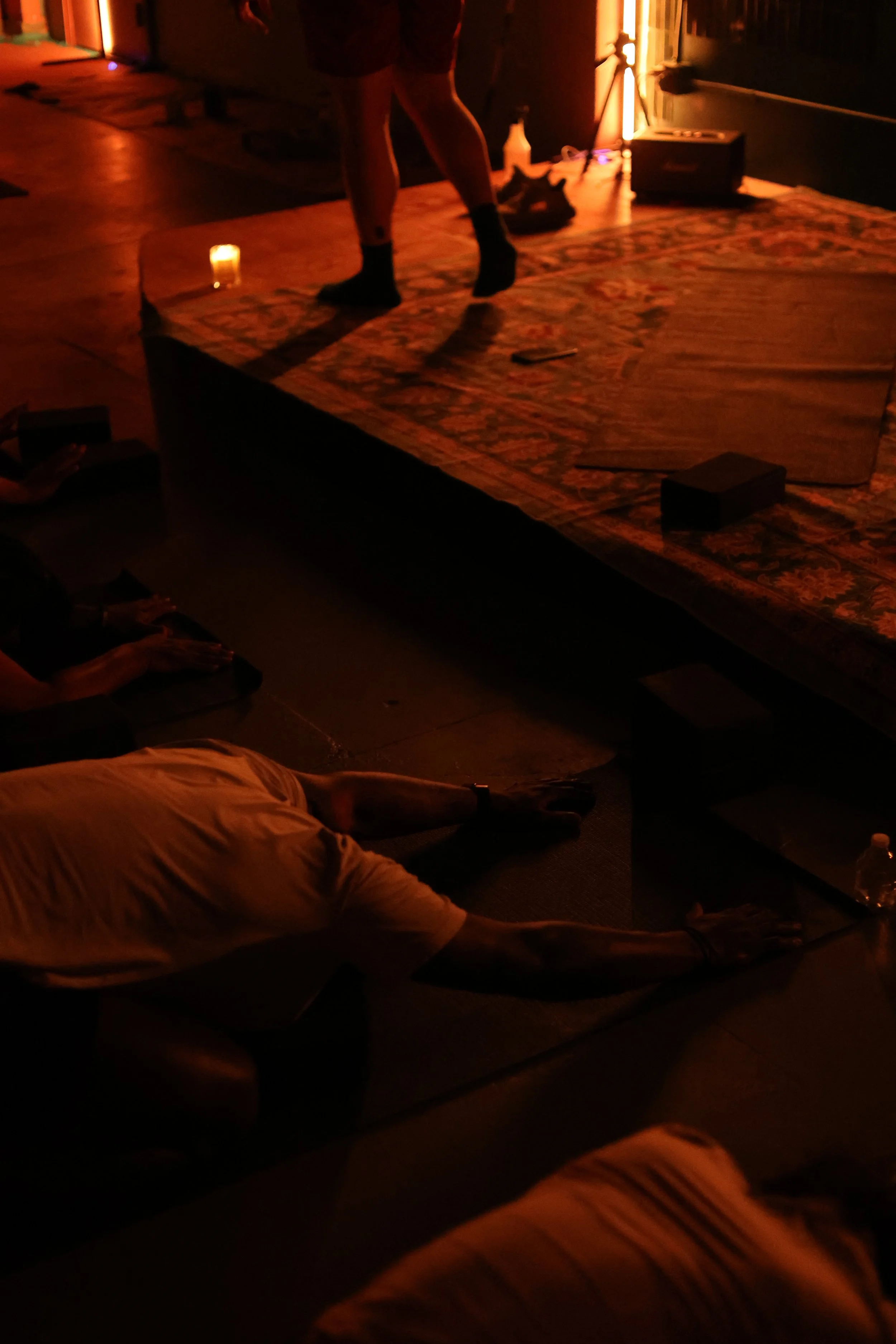 People practicing yoga or stretching on yoga mats in a dimly lit room with warm lighting and a raised platform.