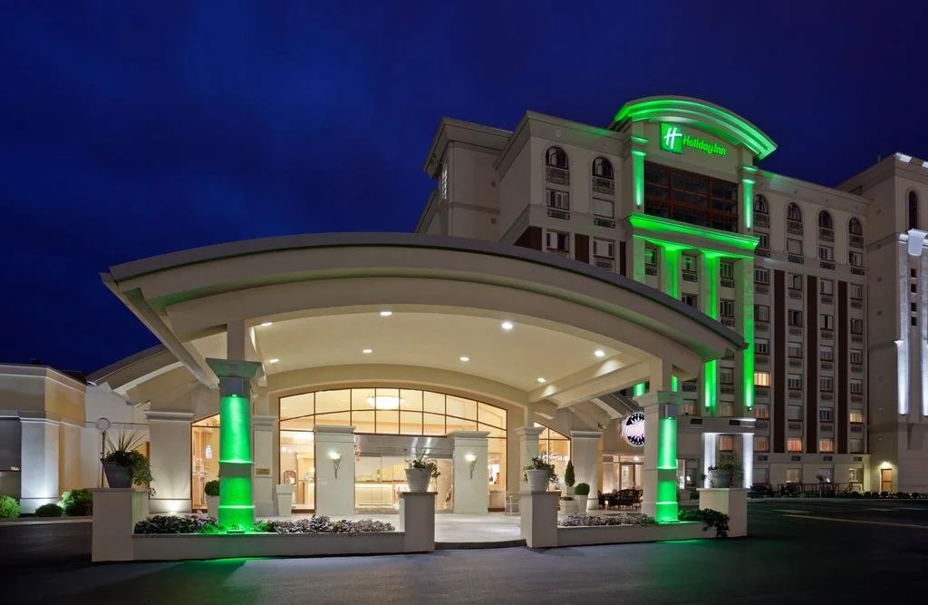 Exterior night view of the Holiday Inn & Suites Parkway Conference Centre in St. Catharines, featuring illuminated green architectural lighting and serving as a major hub for regional corporate events and live streaming.