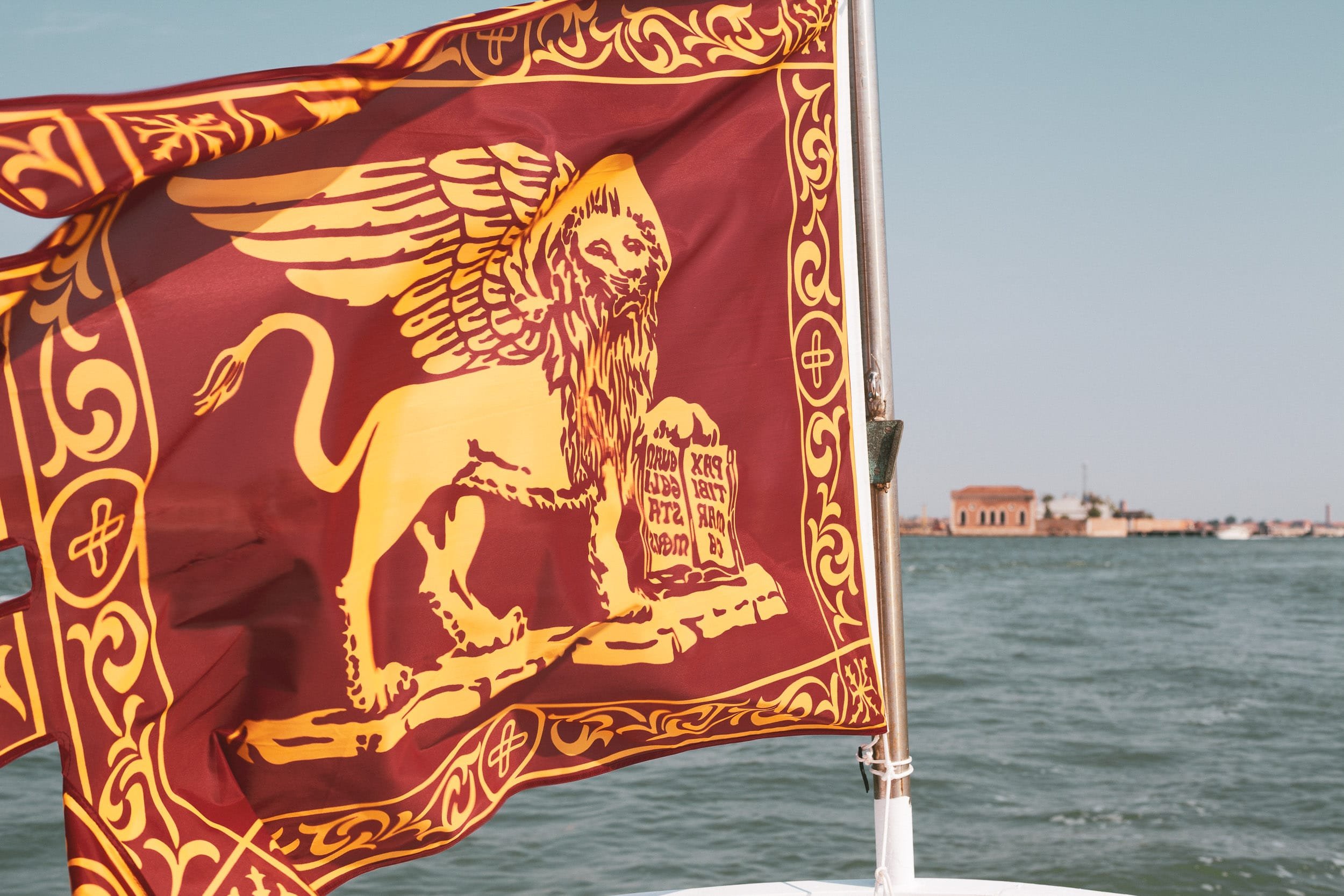 The local winged lion flag red and yellow of Venice floats in the wind with the city in the background.