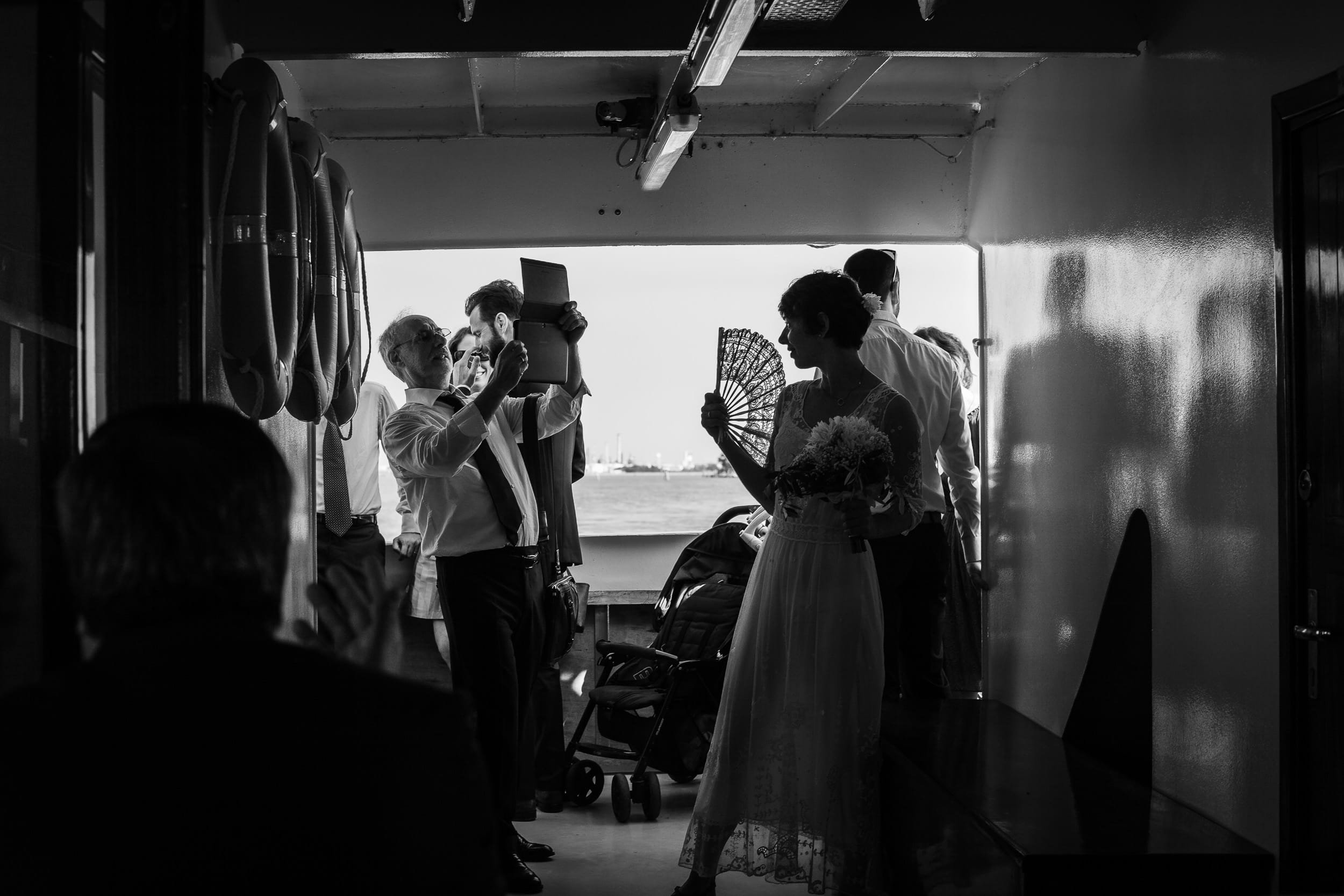 Bride cooling herself off with a fan on a boat in Venice, Italy while a person takes her picture with an Ipad