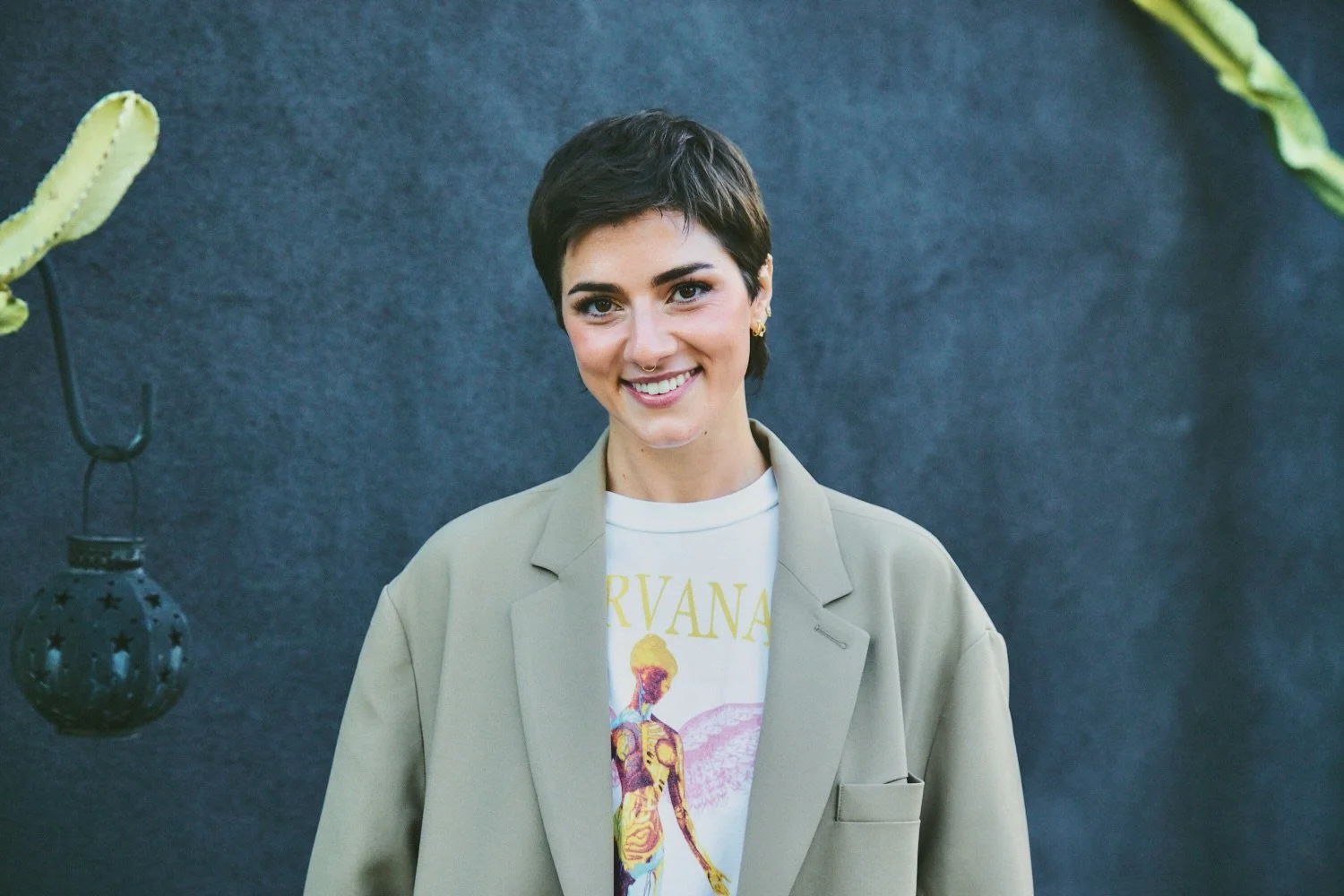A woman with short dark hair, wearing a beige blazer over a Nirvana t-shirt, is smiling at the camera against a dark textured background with hanging decorative objects and green plants.