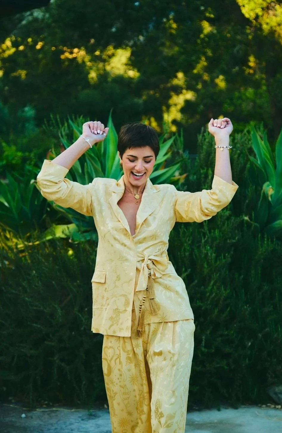 A woman in a yellow patterned suit with rolled-up sleeves, tie-waist, and matching pants, smiling and raising her fists in celebration outdoors with greenery and trees in the background.