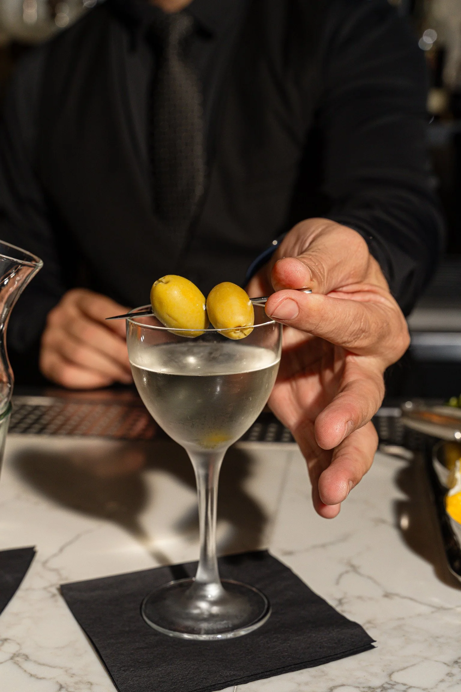 Bartender's hands garnishing a classic martini with olives on a dark marble bar, evoking upscale Chicago steakhouse dining