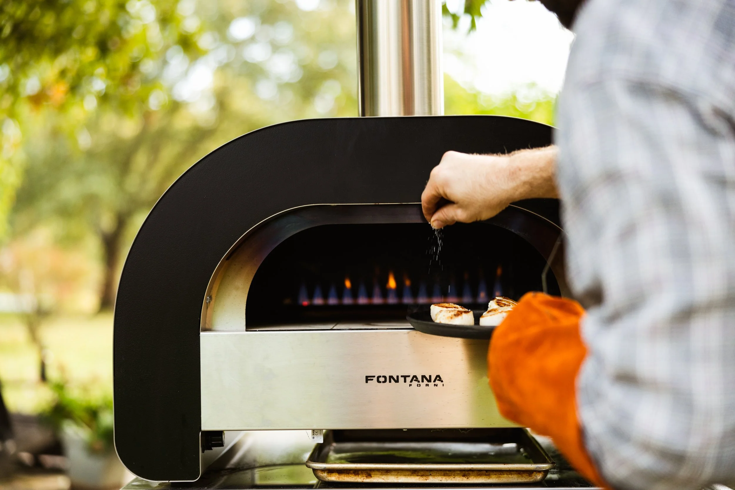 Hands placing food into a sleek black Fontana Forni outdoor pizza oven, highlighting premium artisan cooking equipment
