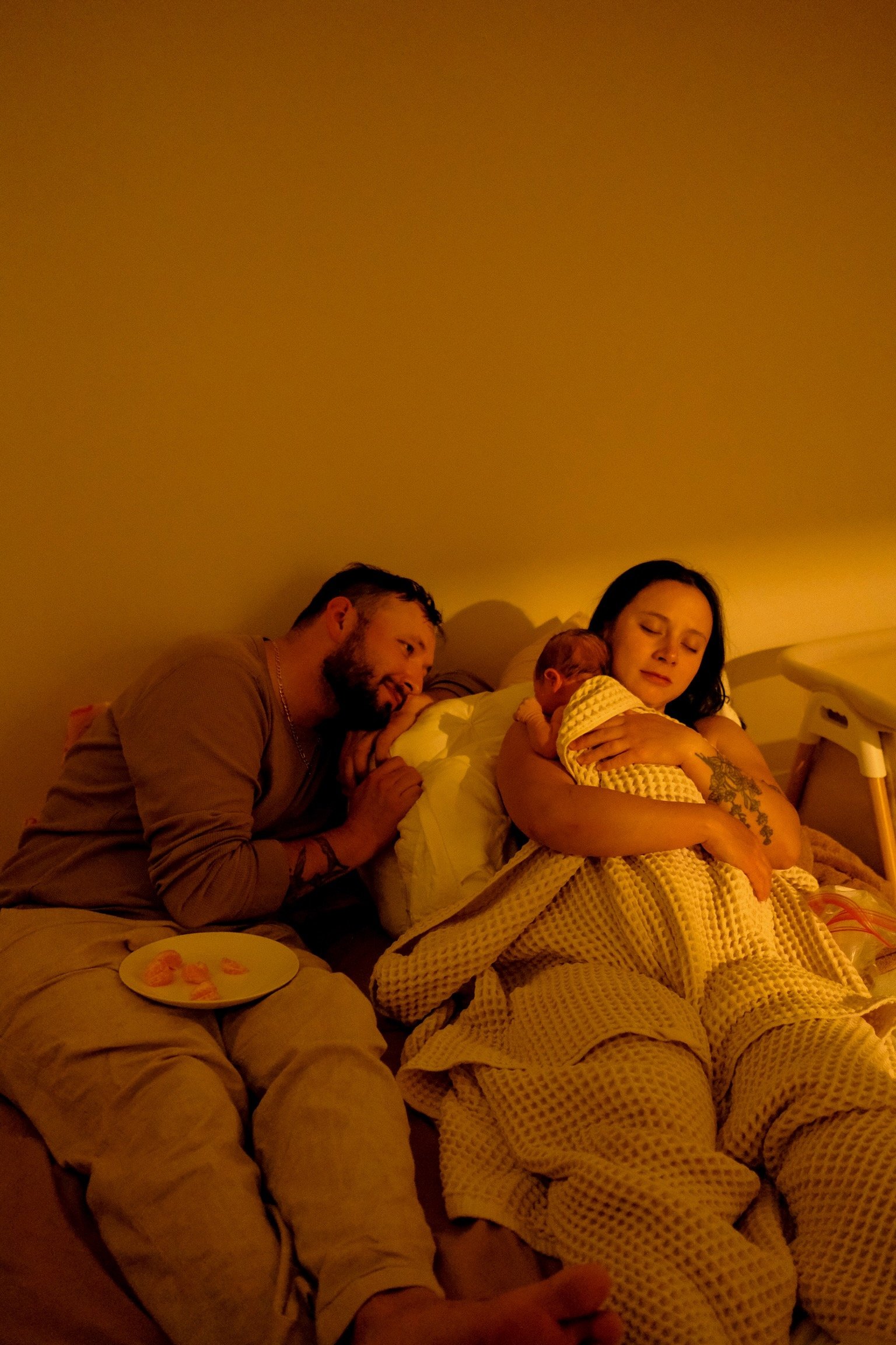 A family of three sitting on a bed in dim warm lighting; a man, woman, and a baby.