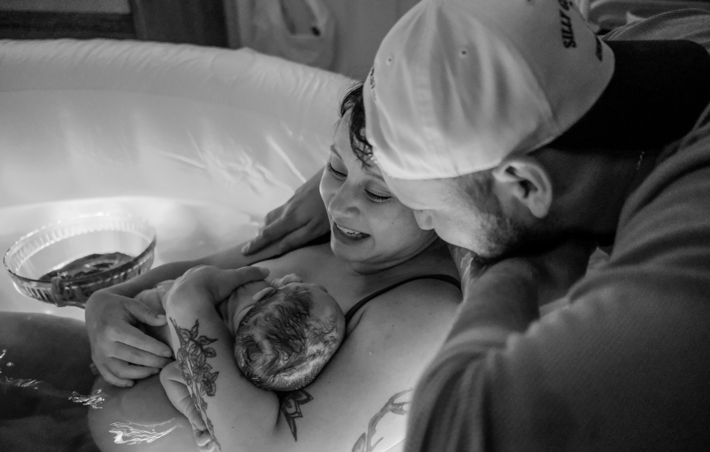 A person holding a newborn baby in a hospital setting, with a woman in the background smiling and looking at the baby, in black and white.