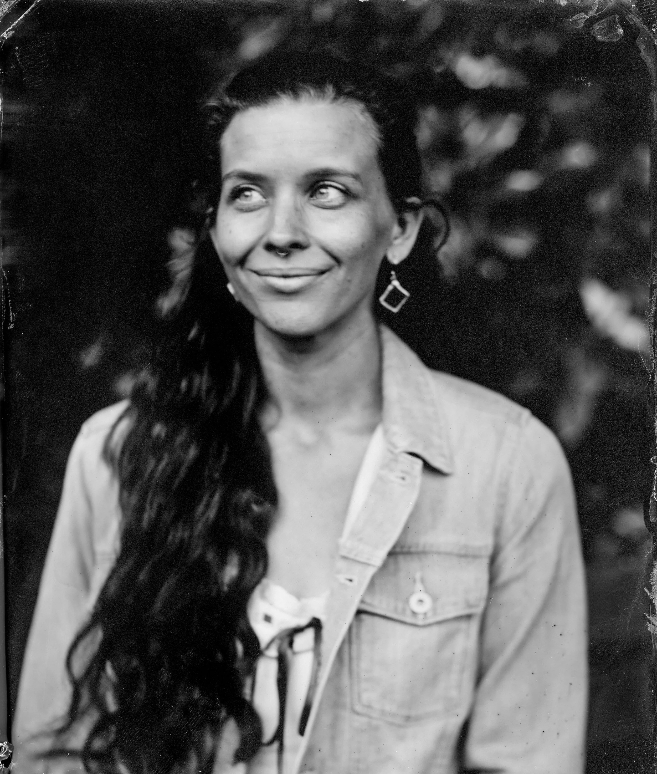 A young woman wearing a white dress and denim jacket, looking off to the side, seated in a garden.
