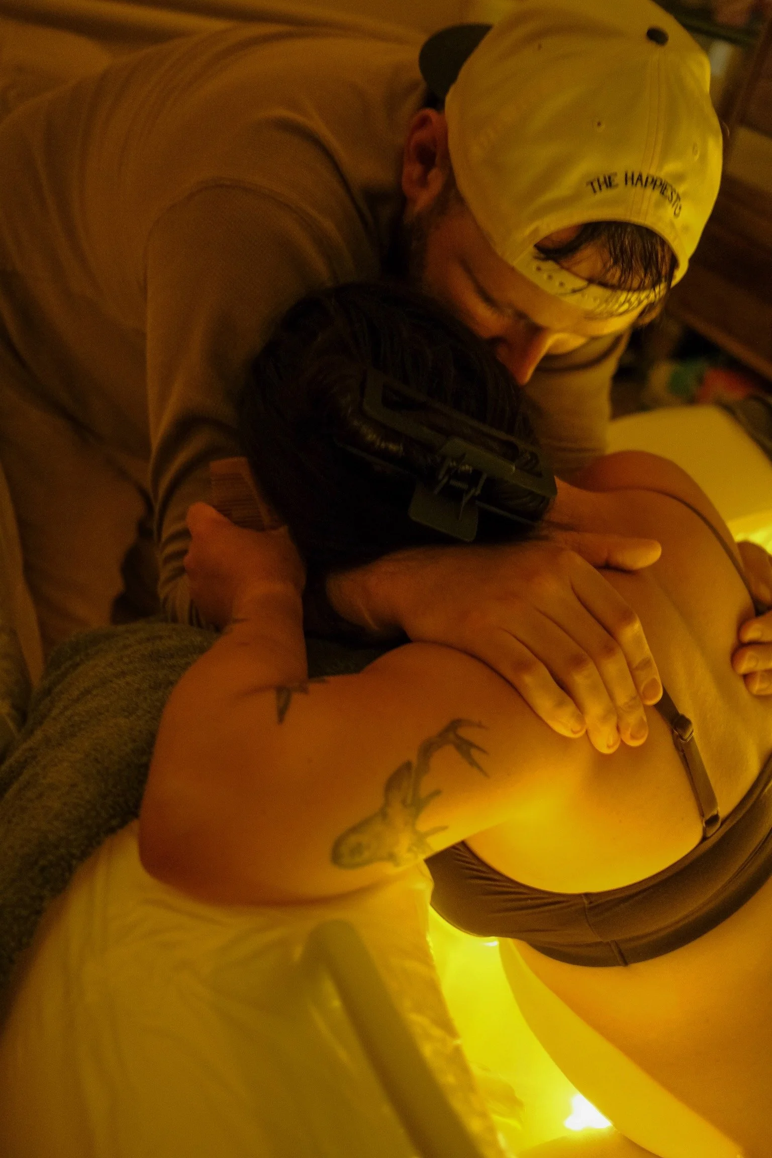A man wearing a yellow cap labeled 'The Happiest' hugging a woman at a party or gathering in a warmly lit room.