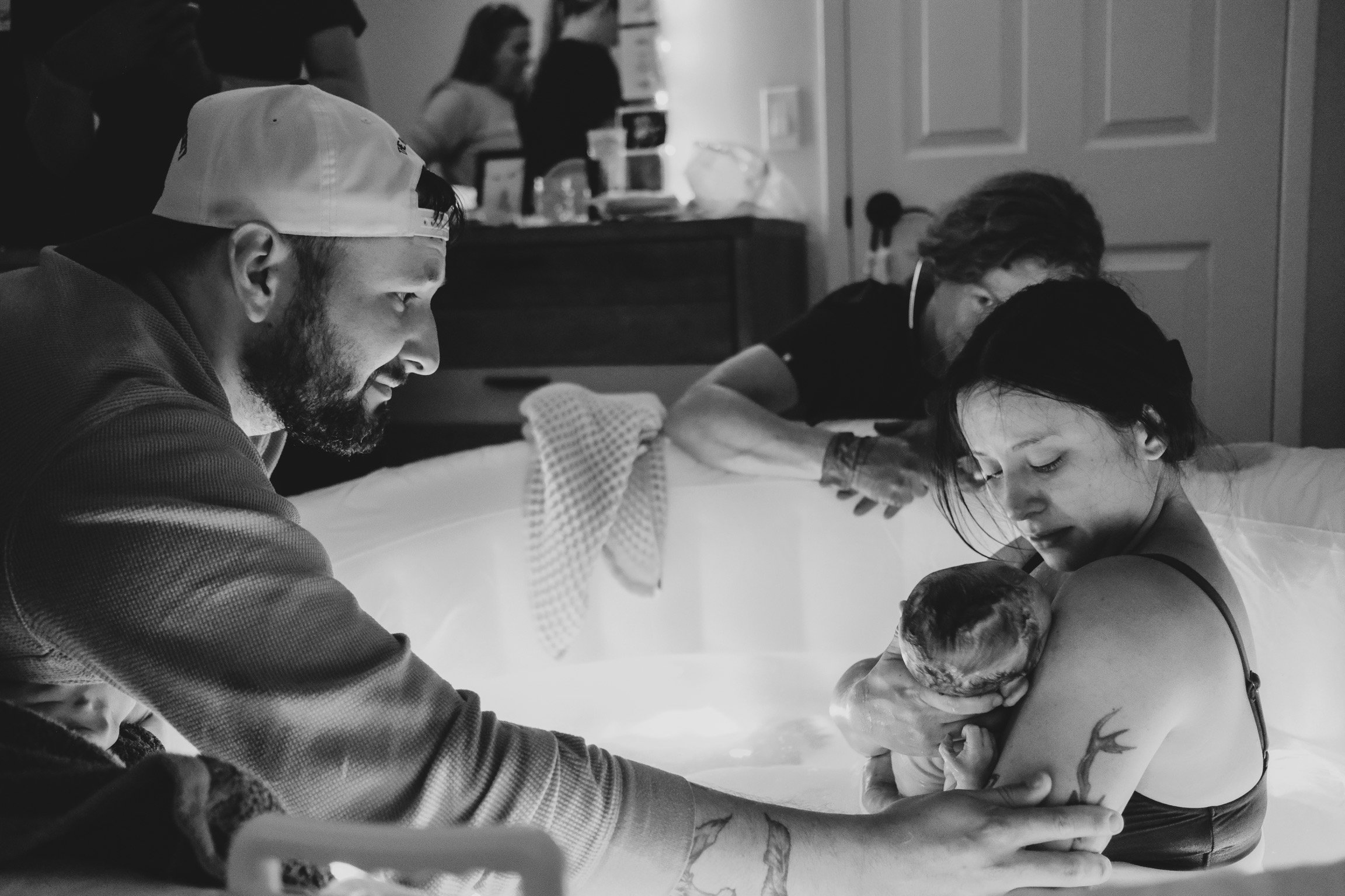 A black and white photo of a man and a woman in a hospital delivering a baby. The man is holding the newborn and the woman is lying in a hospital bed. Medical staff and visitors are in the background.