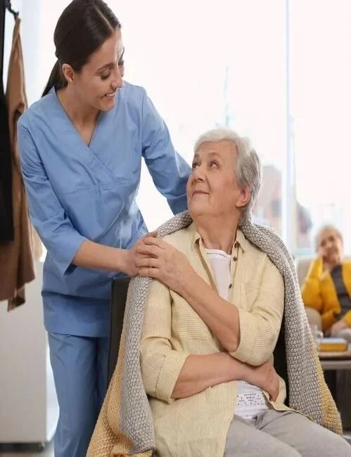 A young female nurse in blue scrubs smiling and talking to an elderly woman with gray hair, who is seated and wearing a beige cardigan, in a bright room with large windows and a blurred person sitting in the background.