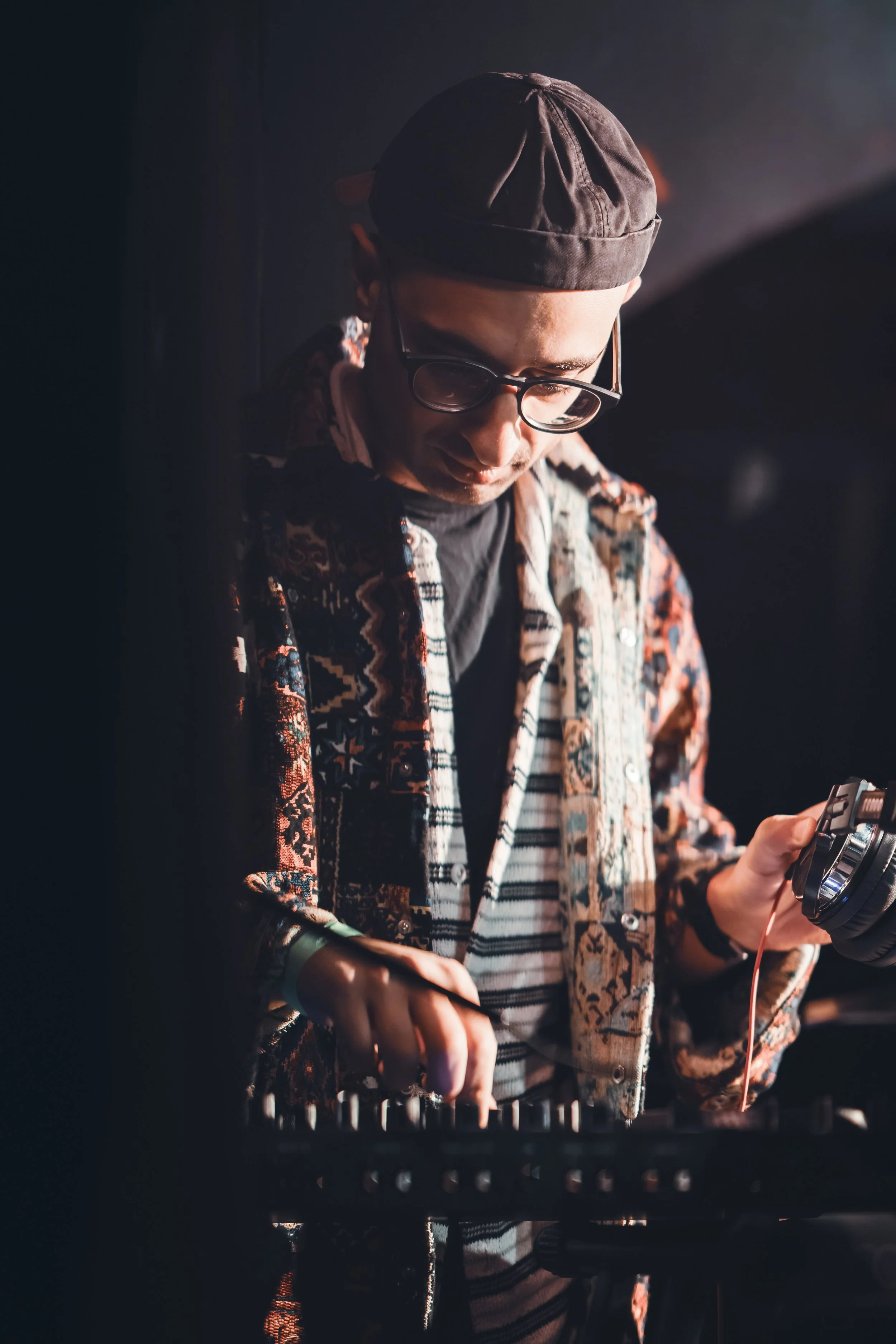 DJ wearing glasses, patterned shirt, and cap, adjusting equipment in a dimly lit setting.