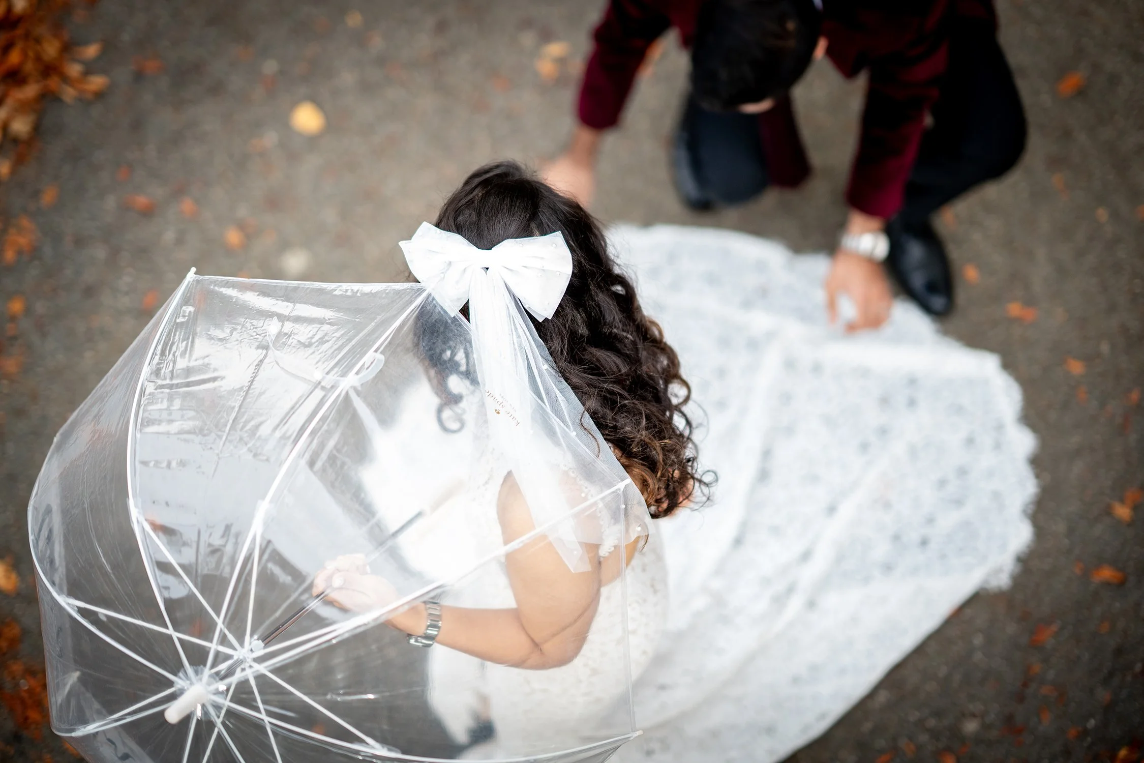 A bride dressed in a white wedding gown holding a transparent umbrella with a white bow, standing on a paved pathway with fallen leaves, viewed from above, with a man kneeling nearby.