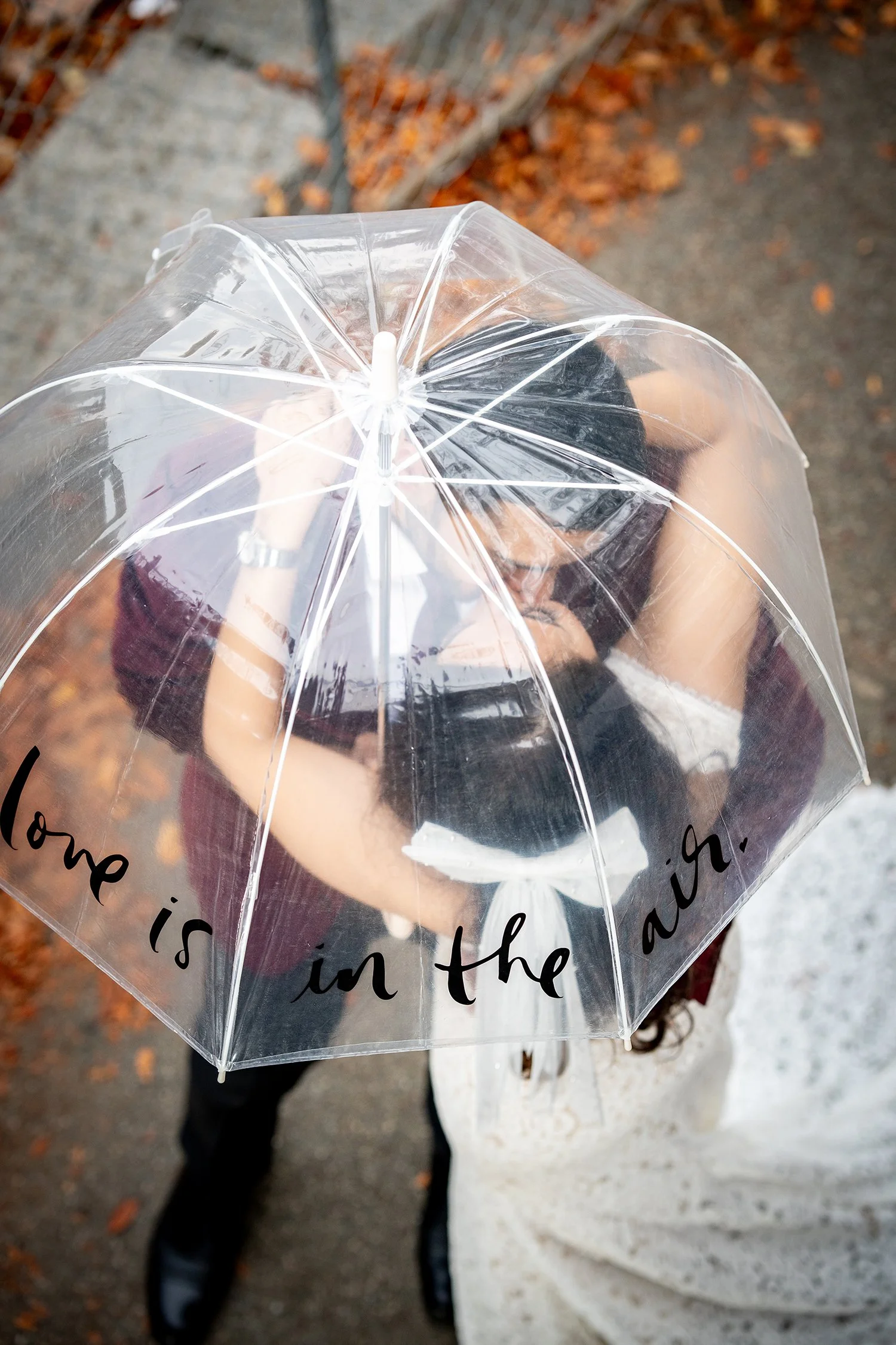 Two women kissing beneath a transparent umbrella with the words 'Home is in the air' written on it, on an outdoor path with fallen autumn leaves.