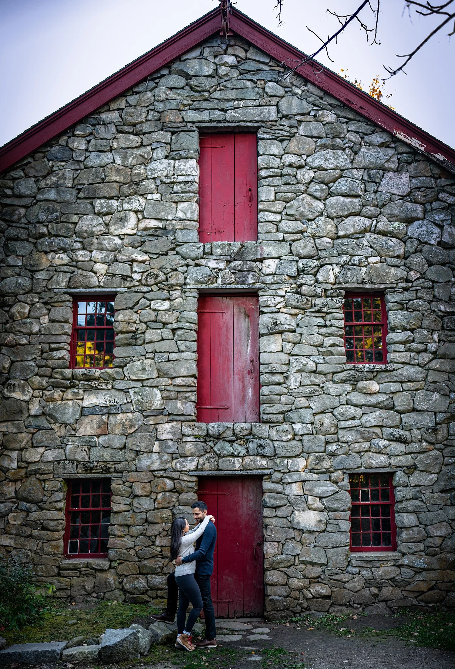 A couple is embracing and dancing in front of a rustic stone house with red doors and windows, outdoors during daylight.