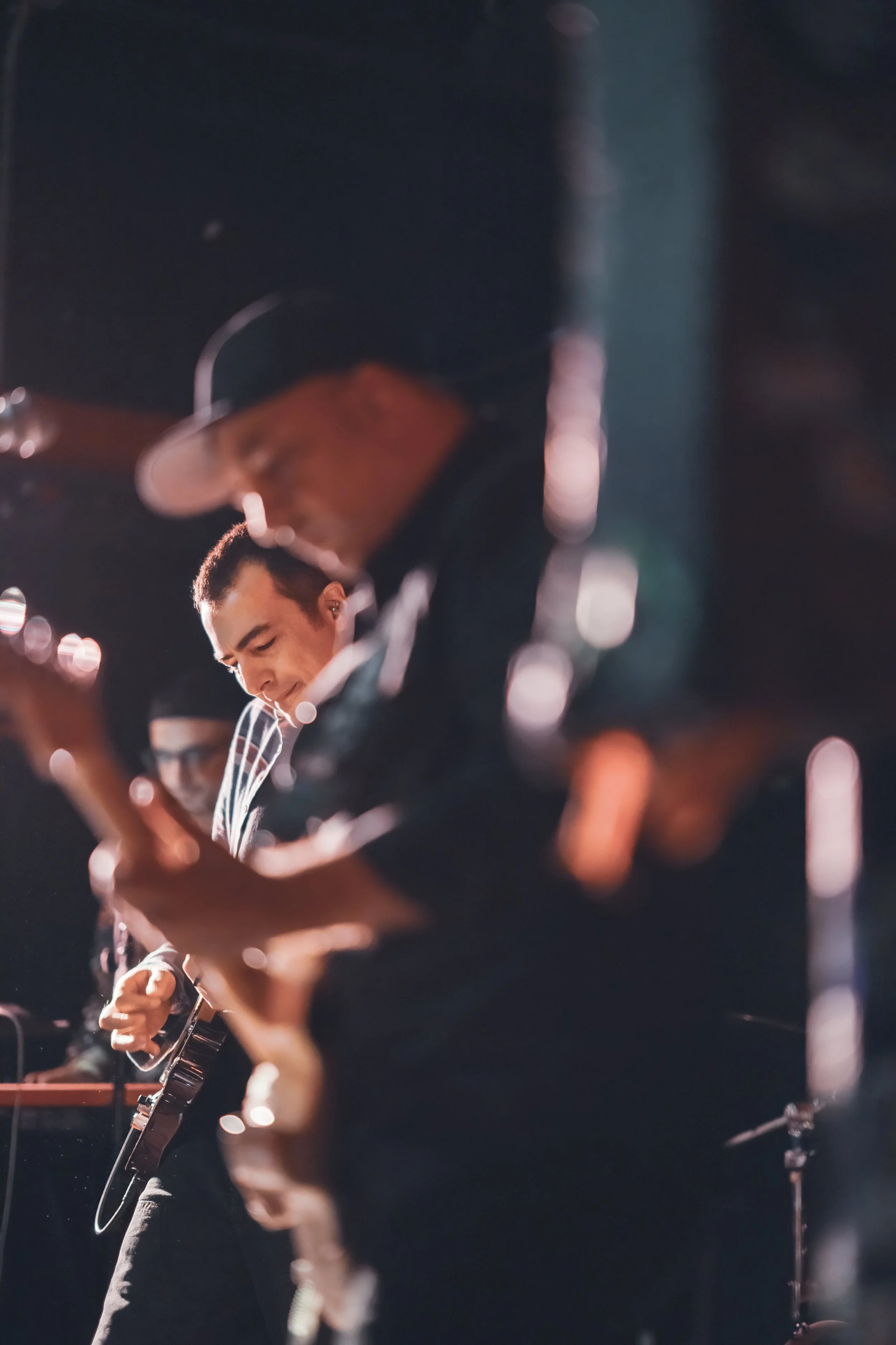 Musicians playing guitars during a live performance, with a focus on the man in the foreground and others slightly blurred in the background.