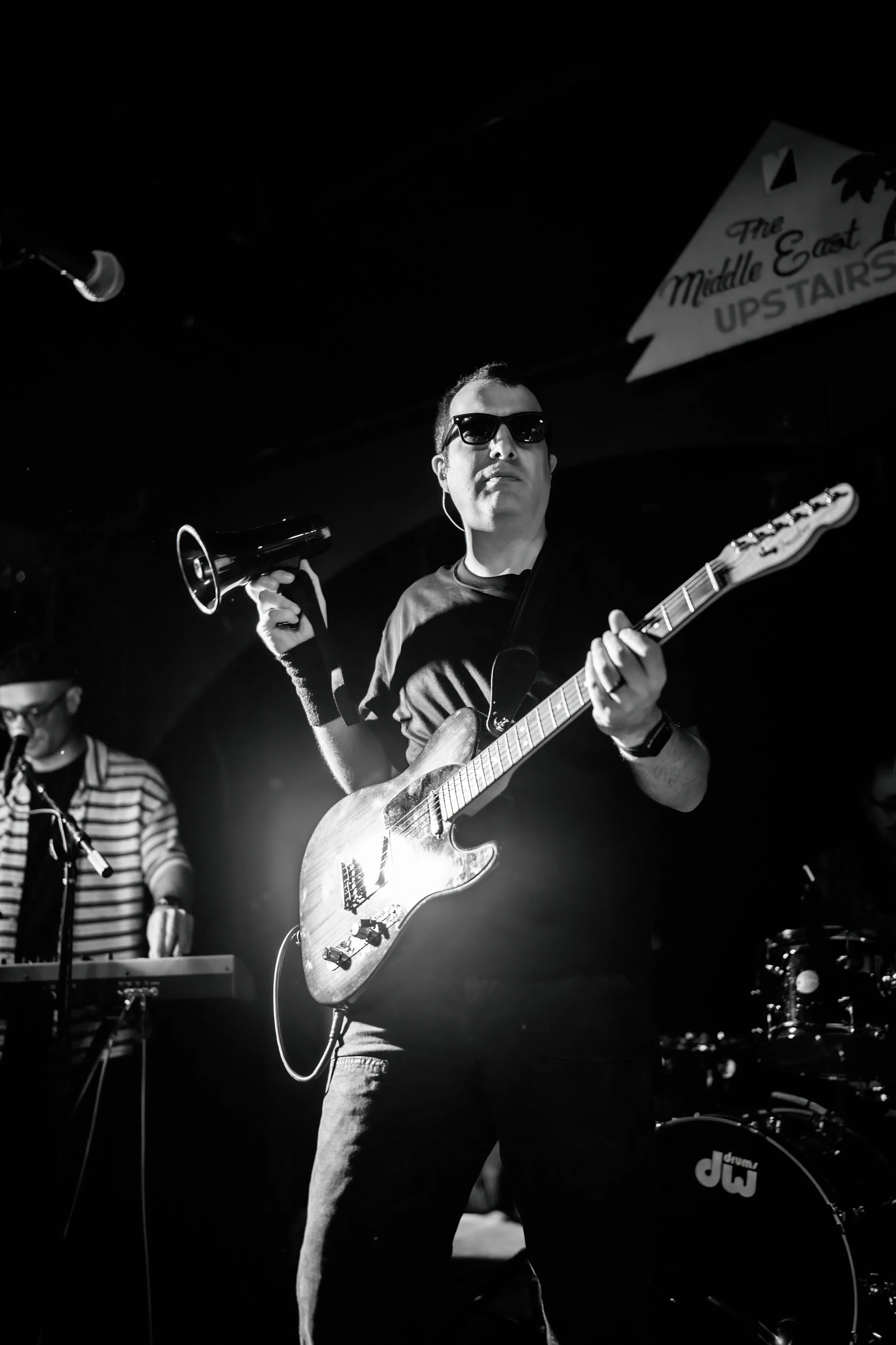 A musician wearing sunglasses and holding an electric guitar and a megaphone on stage during a performance, with a keyboard player in the background.
