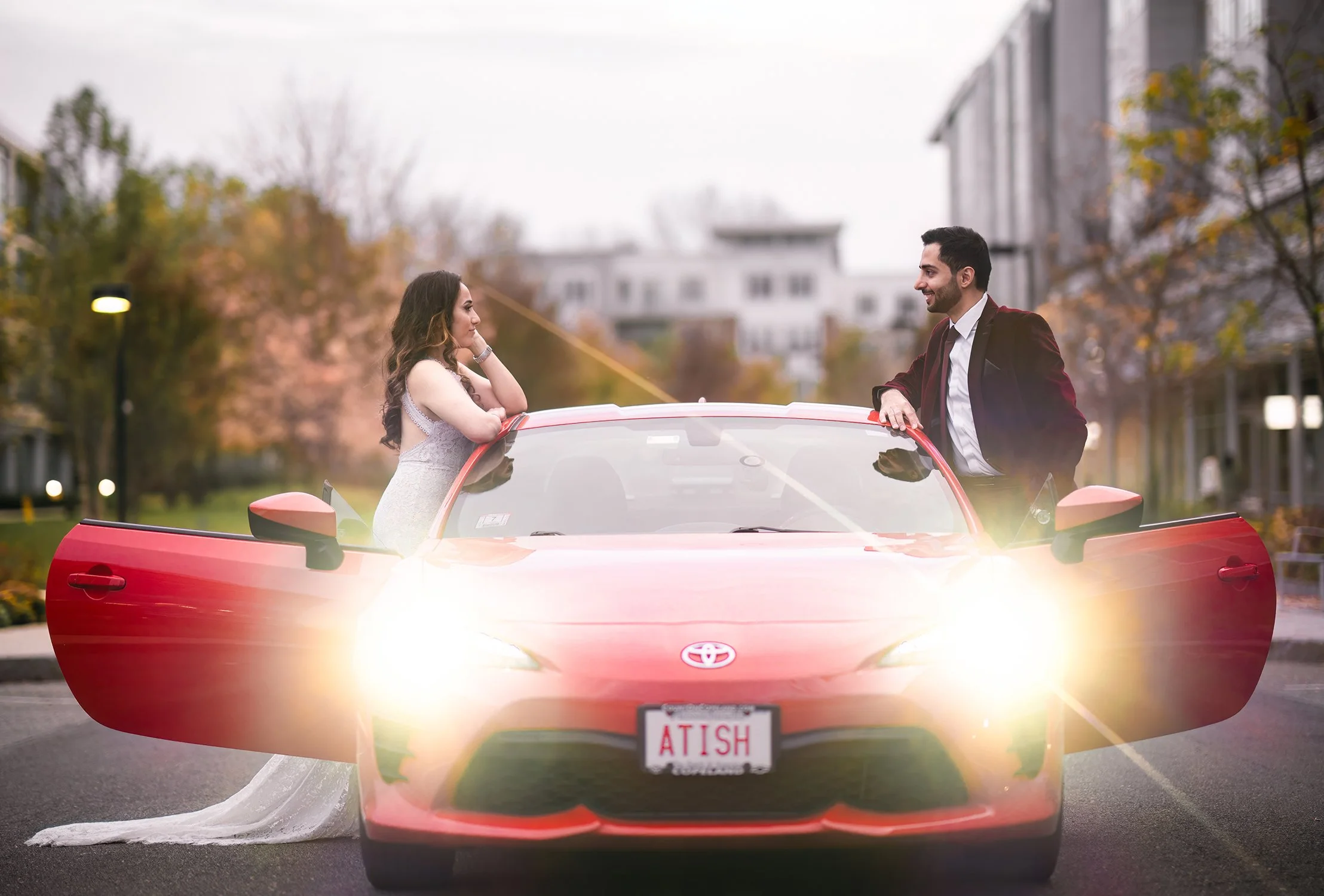 A bride and groom standing on opposite sides of a red Toyota convertible with open doors, in an urban park setting during dusk, with the groom smiling and the bride looking pensive.