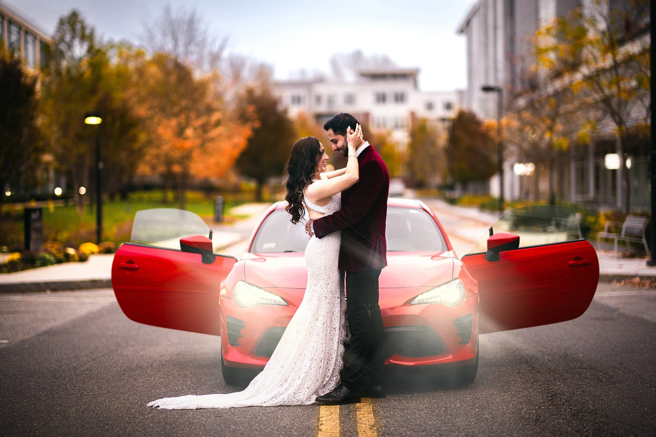 A bride and groom kissing in front of a red sports car with open doors, on a city street with autumn trees and buildings in the background.