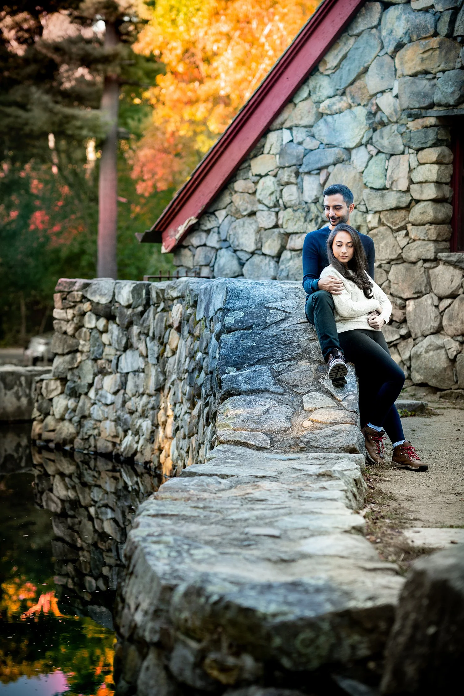 A man and woman sitting on a stone wall beside a small pond with a stone building in the background during autumn, with colorful fall foliage.