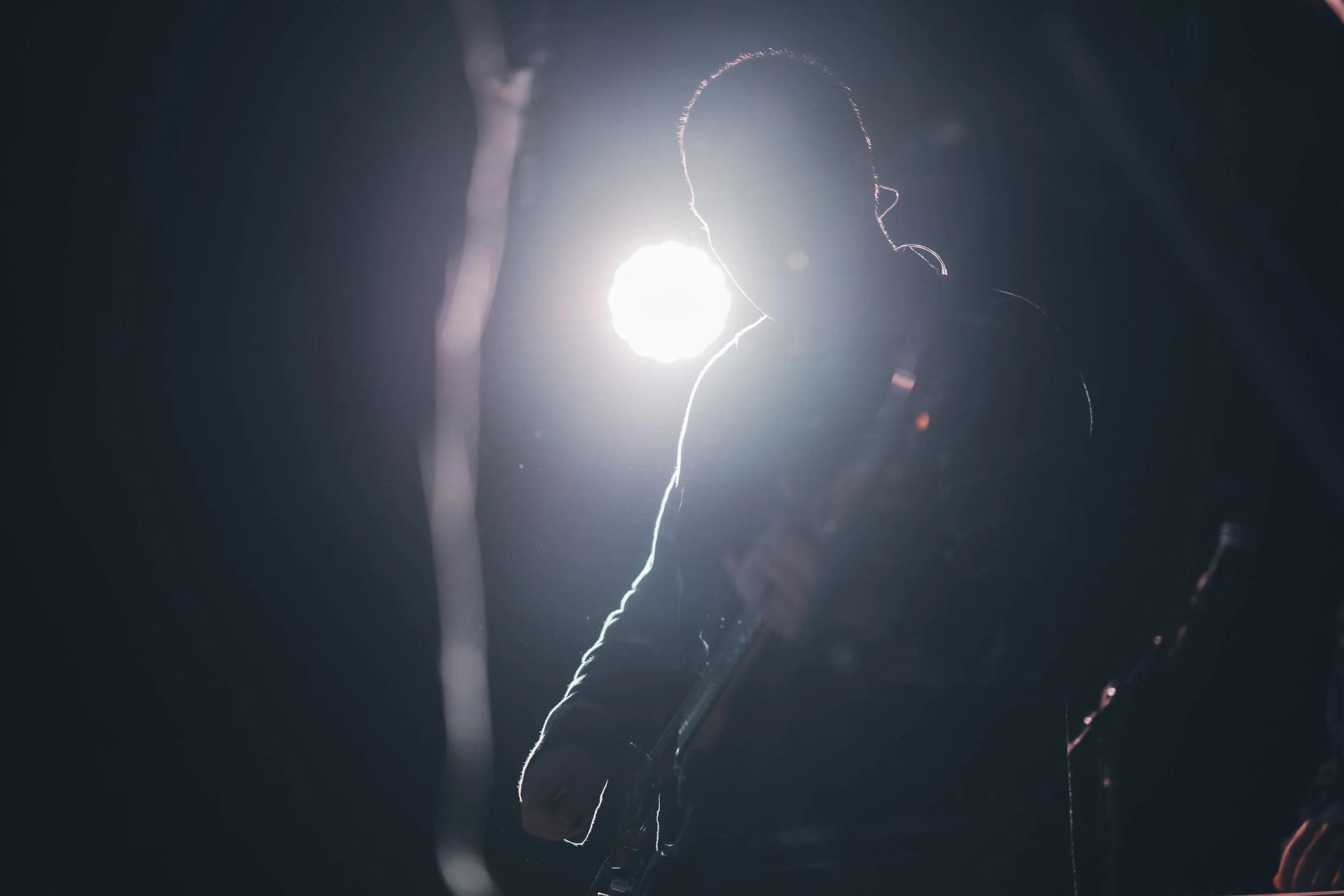 Silhouette of a person holding a guitar, backlit by a bright light, in a dark environment.