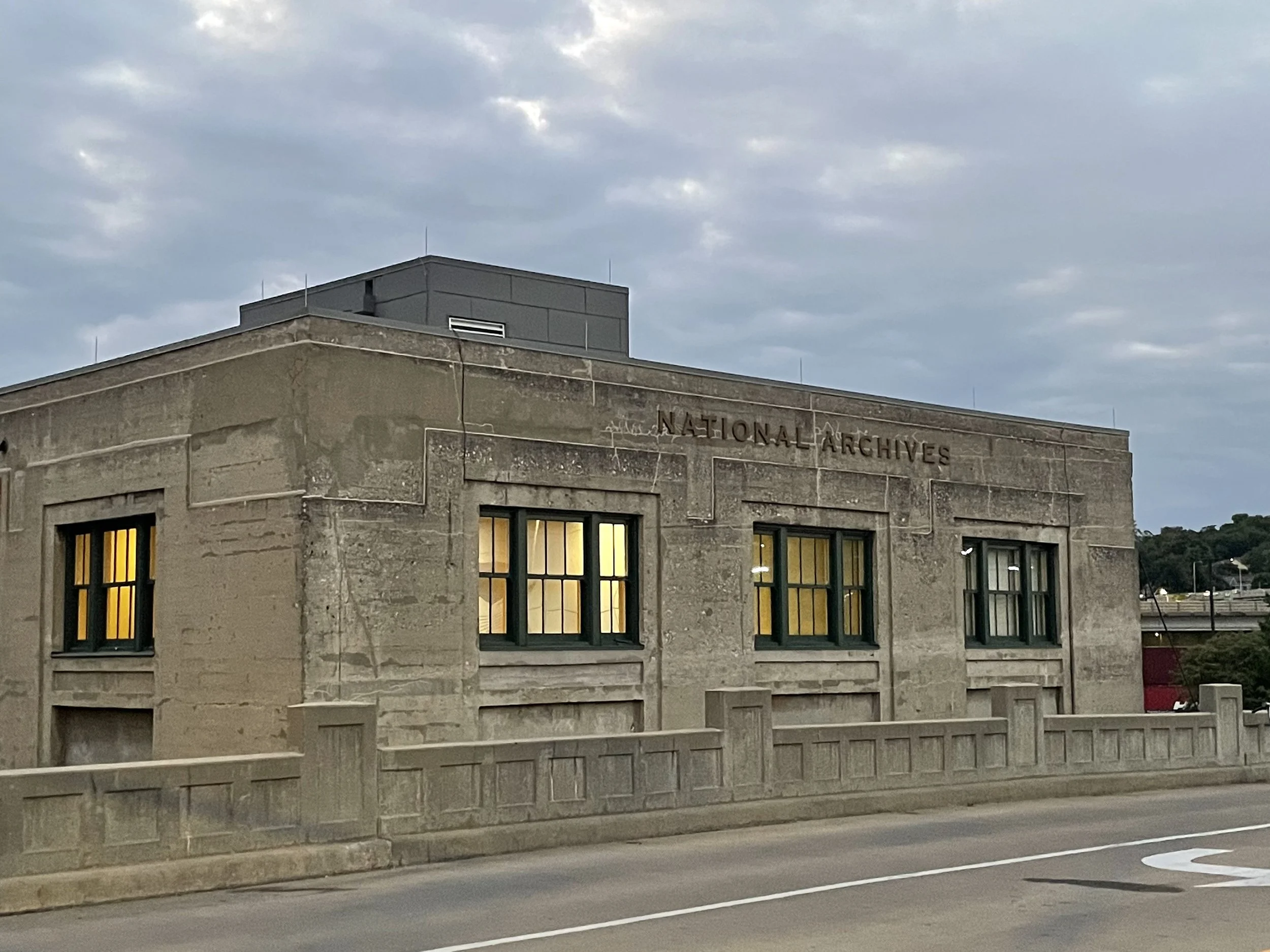 The image shows the National Archives building in Kansas City, situated near a road with street markings. The building has large windows with some lights on inside, and the sky above is cloudy, indicating an overcast day.