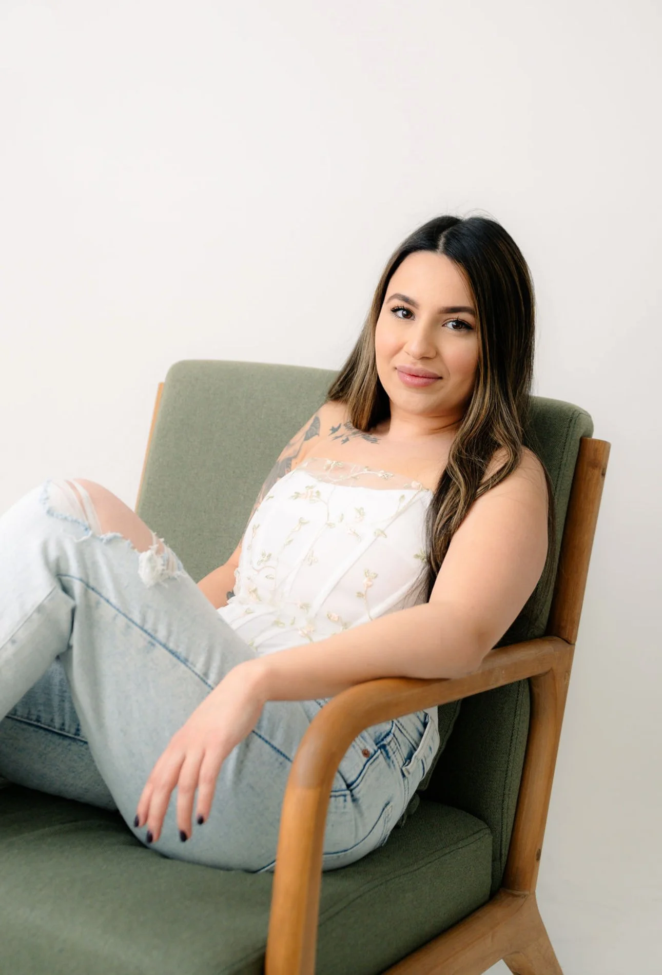 Young woman with long dark hair sitting on a green and wooden chair, wearing a white embroidered top and ripped jeans, looking at the camera with a slight smile.