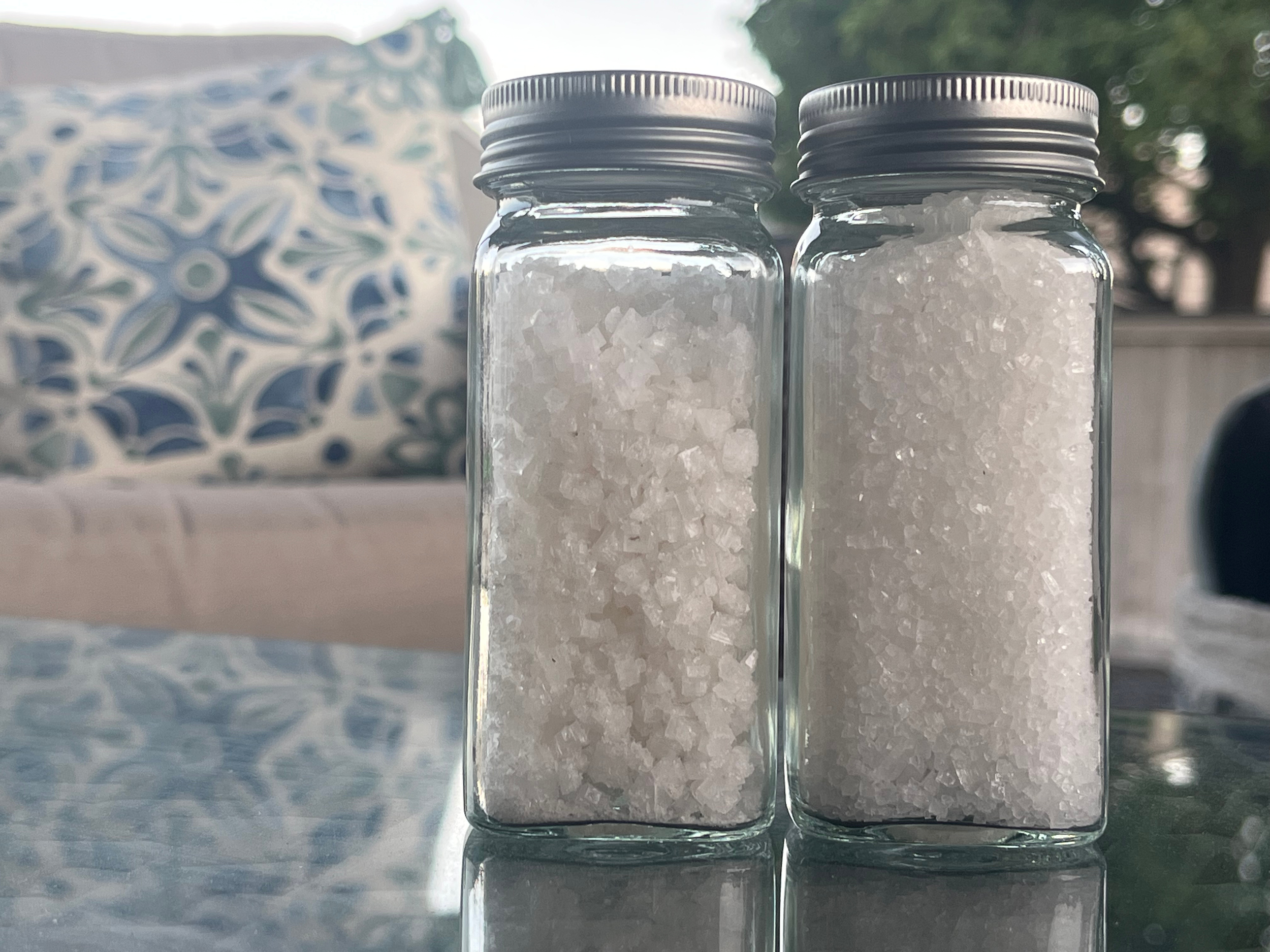 Two glass jars filled with salt placed on a reflective surface outdoors, with a patterned cushion and trees in the background.