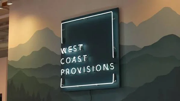 Neon sign reading 'West Coast Provisions' mounted on a mountain-themed mural wall.