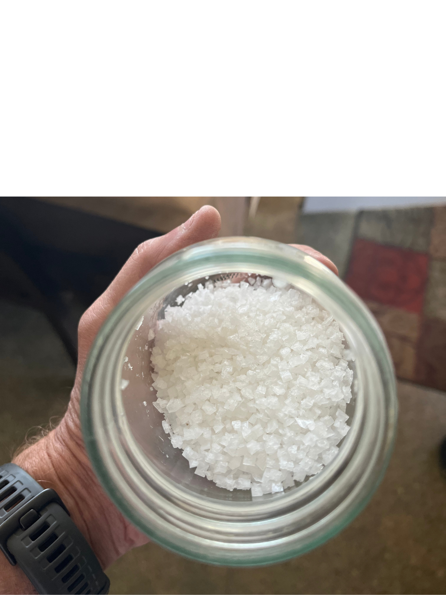 A hand holding an open jar of sea salt with large white flakes, against a background of a tiled floor and wooden surface.