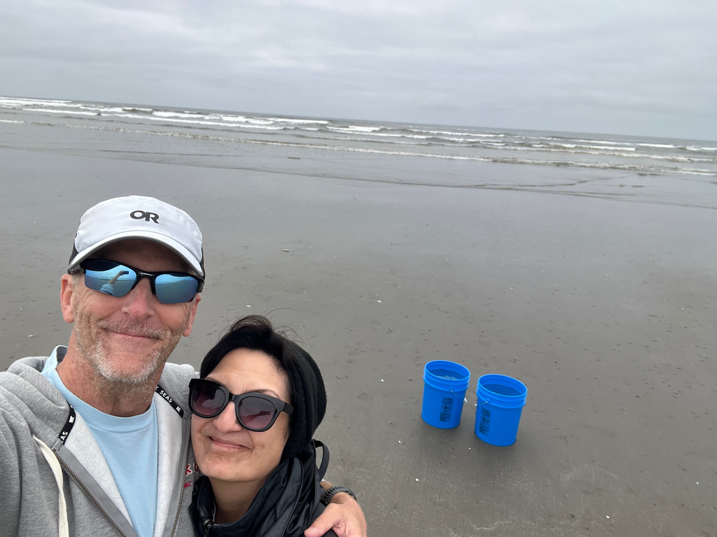 A smiling man and woman taking a selfie on a cloudy beach, with two blue buckets on the sand behind them and the ocean in the background.
