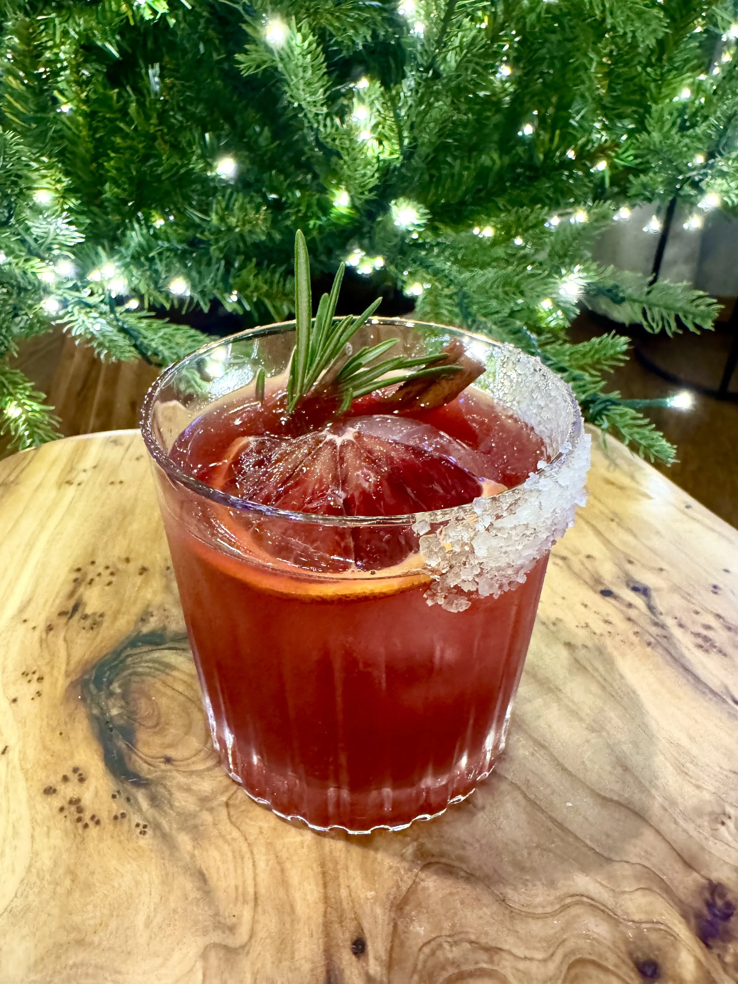 A festive cocktail garnished with rosemary and blood orange slices, with a salted rim on the glass, set on a wooden surface in front of a decorated Christmas tree with twinkling lights.