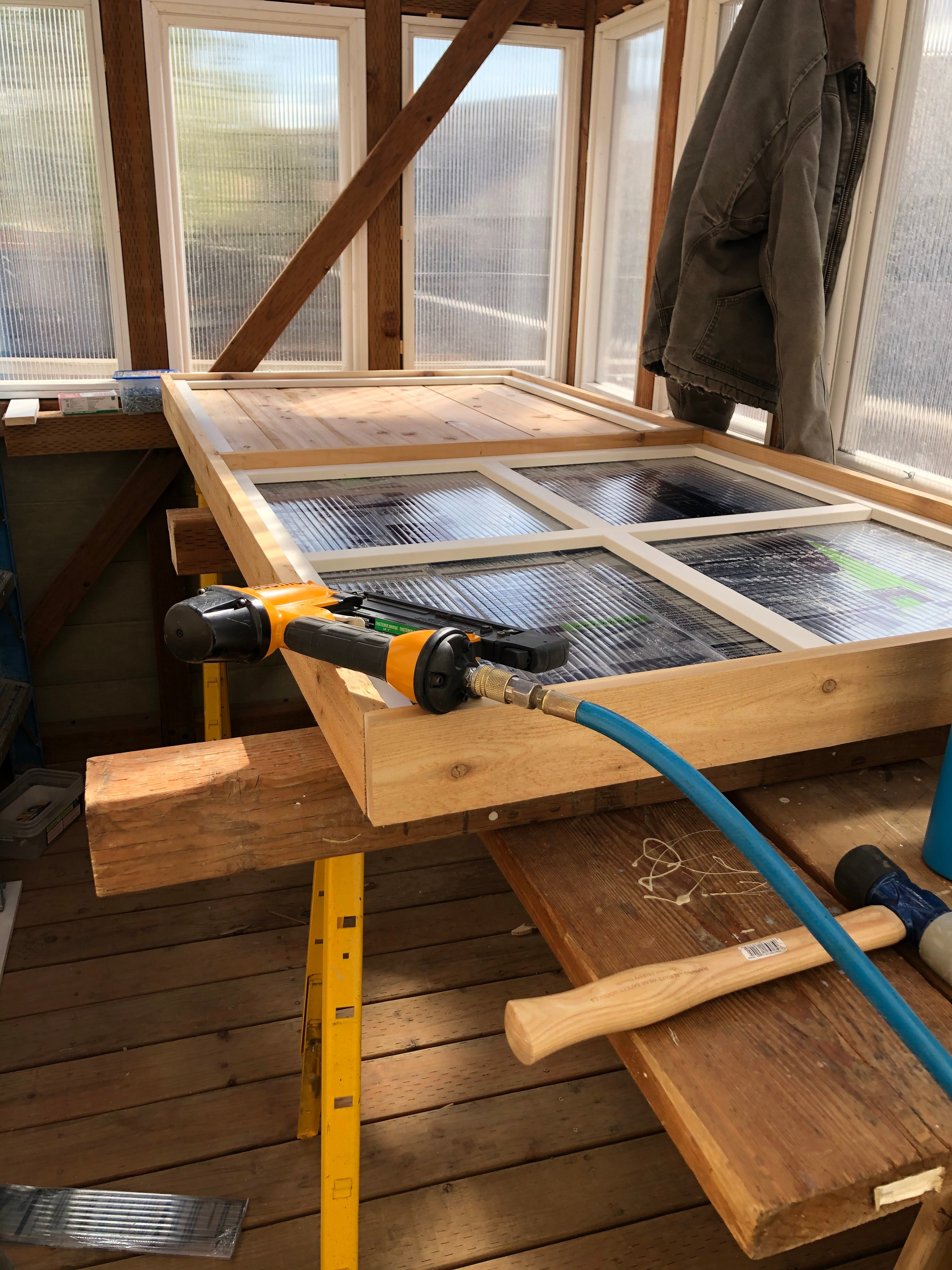 Inside a woodworking workshop, a wooden frame with glass panels is under construction on a workbench, with a hammer and pneumatic nail gun nearby, and a jacket hung inside an enclosure with windows.