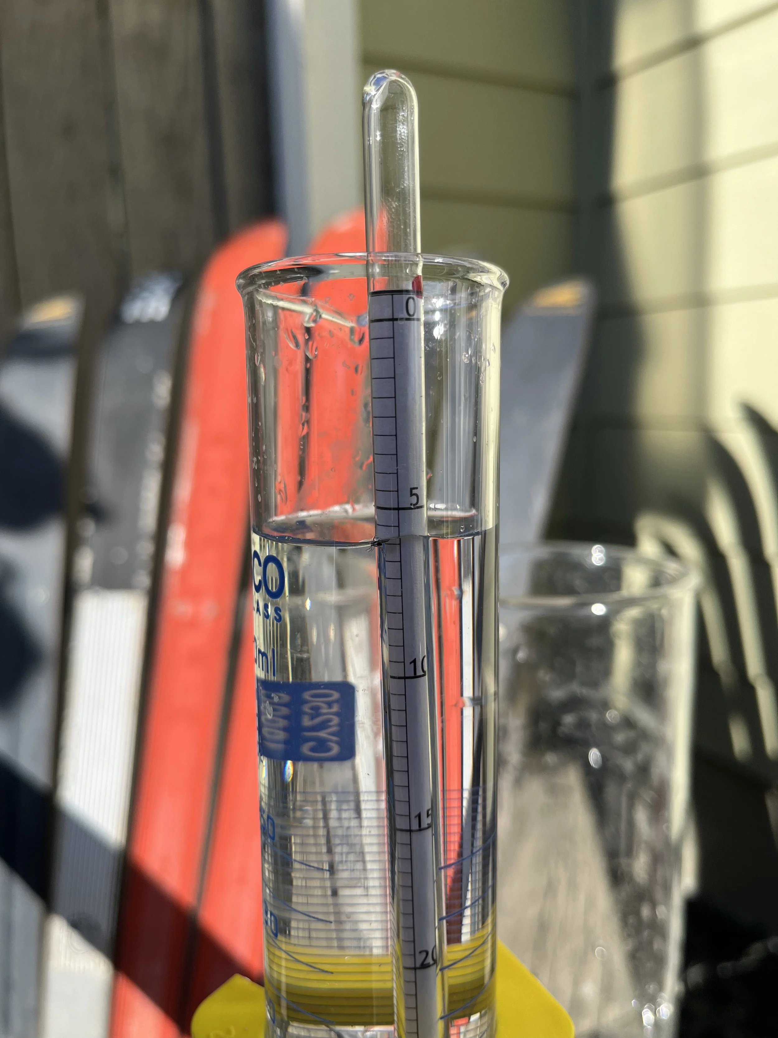 A glass graduated cylinder filled with water and a thermometer resting inside, positioned outdoors near chairs and a building wall.