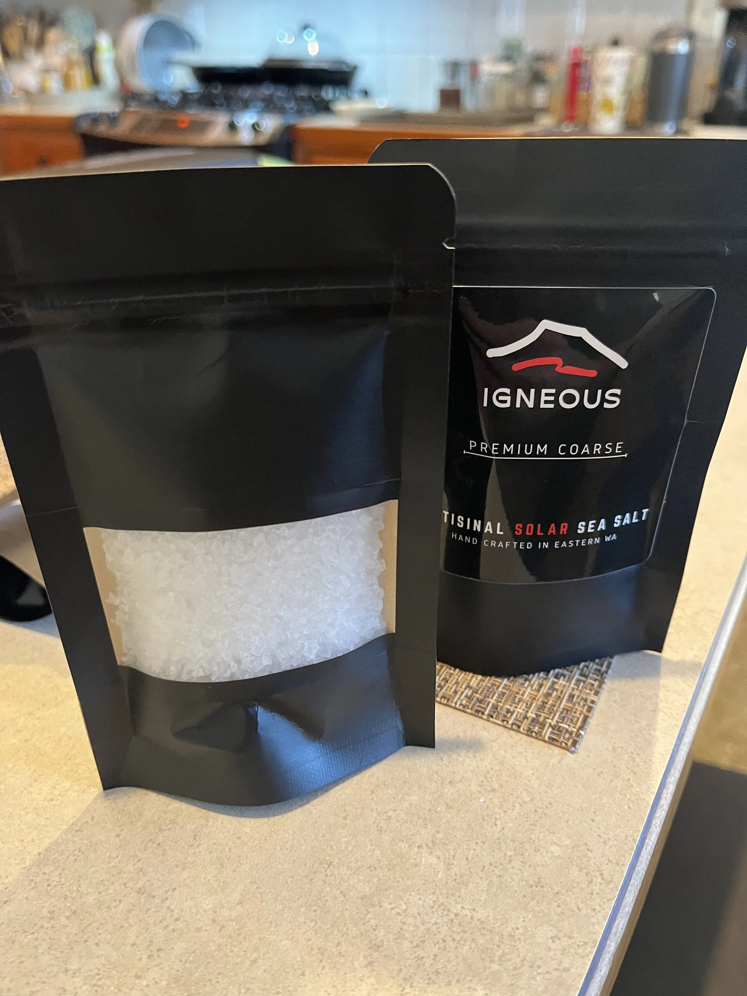 Two Kraft packages of Igneous Salt black packages of sea salt on a beige countertop, one showing coarse white salt crystals and the other labeled 'IGNEOUS Premium Coarse Artisanal Solar Sea Salt'.