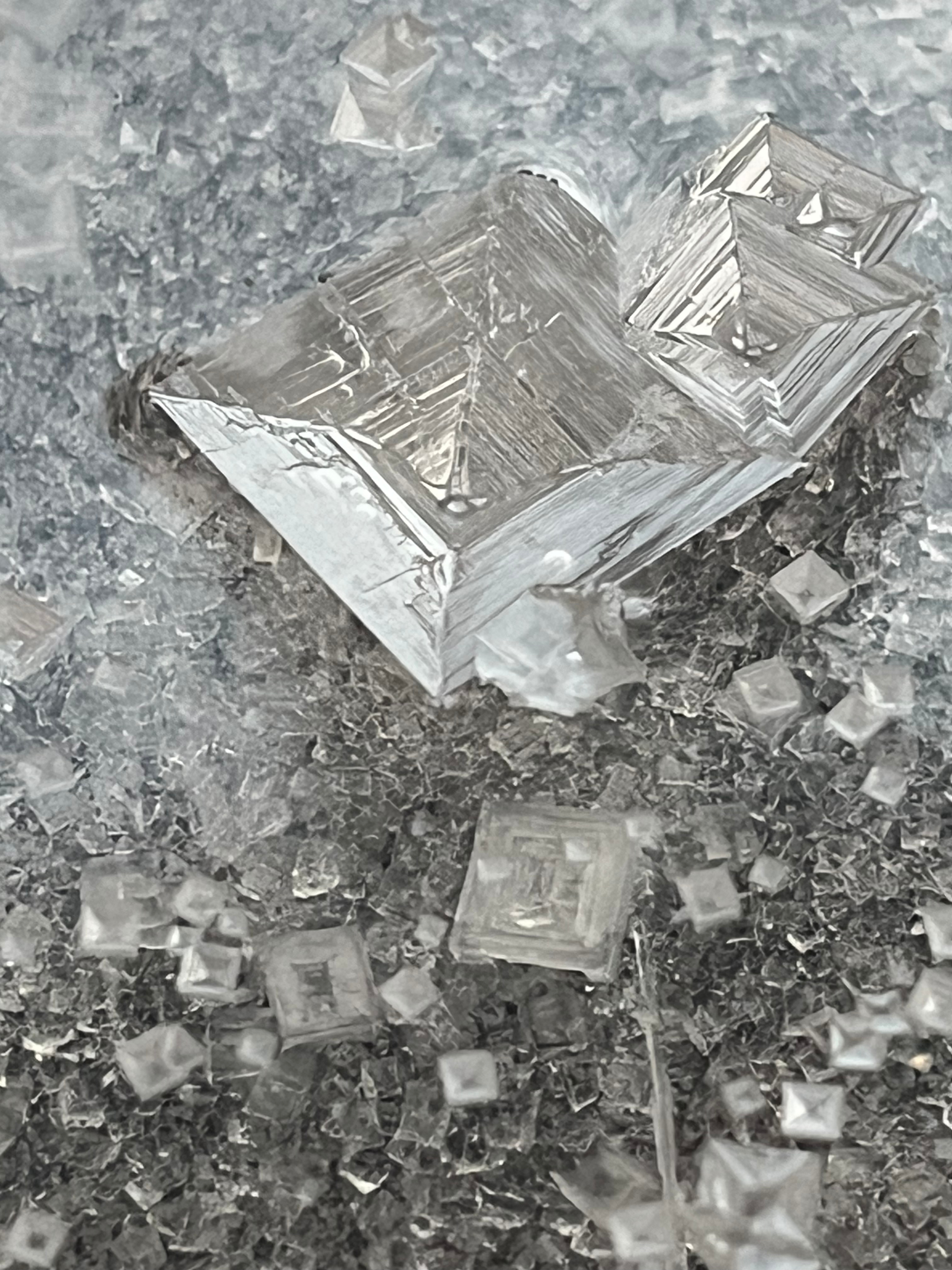 Close-up photograph of a silver, metallic, geometric-shaped object lying on a textured, reflective surface with scattered small, square particles.