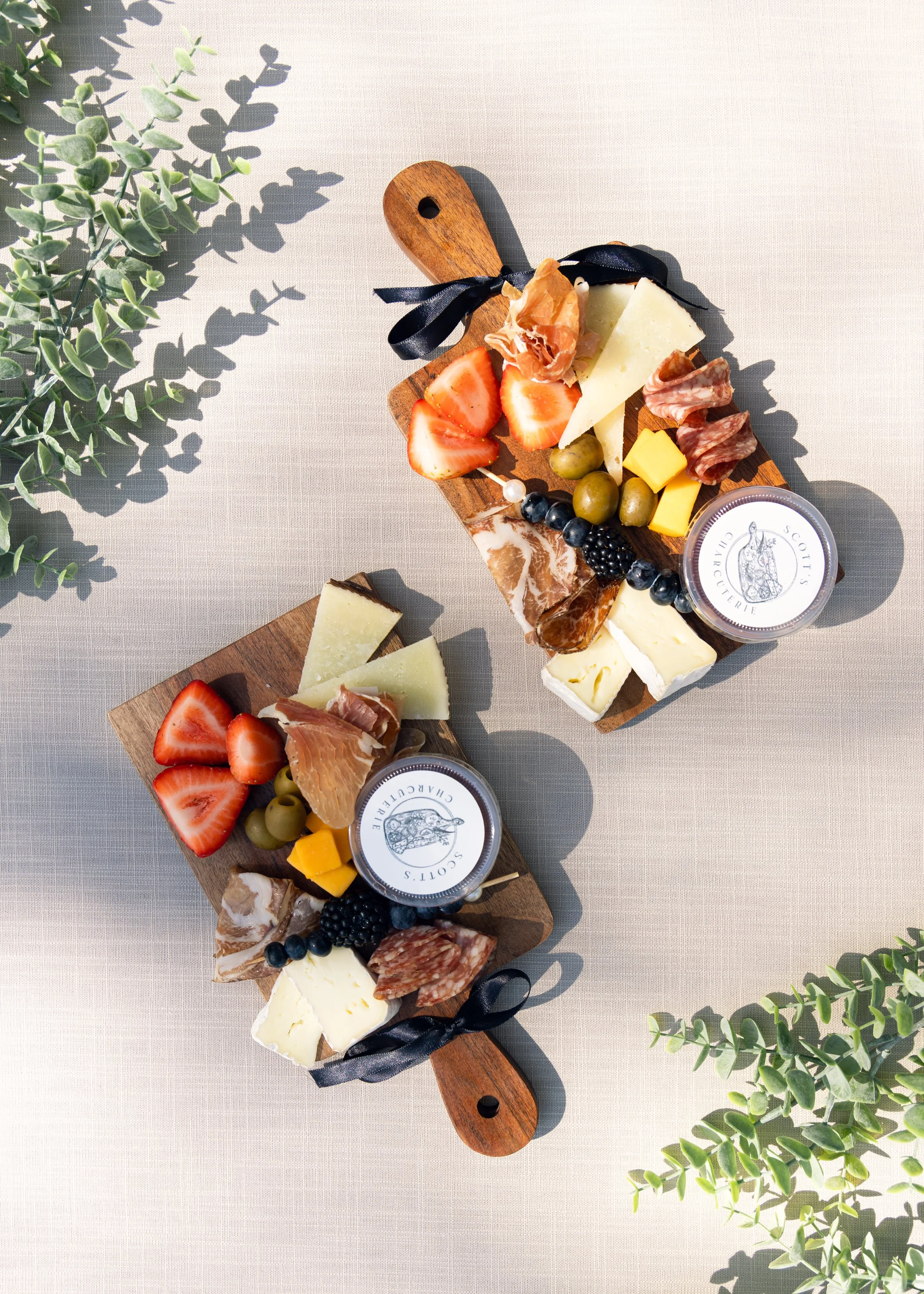 Two individual charcuterie boards with cheese, strawberries, olives, salami, prosciutto, blackberries, crackers, and jam on a light table. 