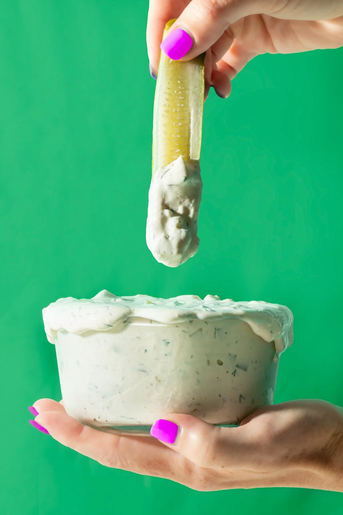 Person holding a bowl of creamy dip with a pickle spear dipped in it, against a green background.