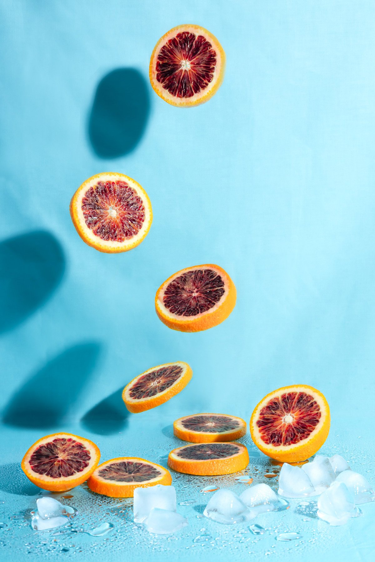 Slices of blood orange falling onto ice cubes on a light blue surface with a matching background, creating a refreshing summer vibe.