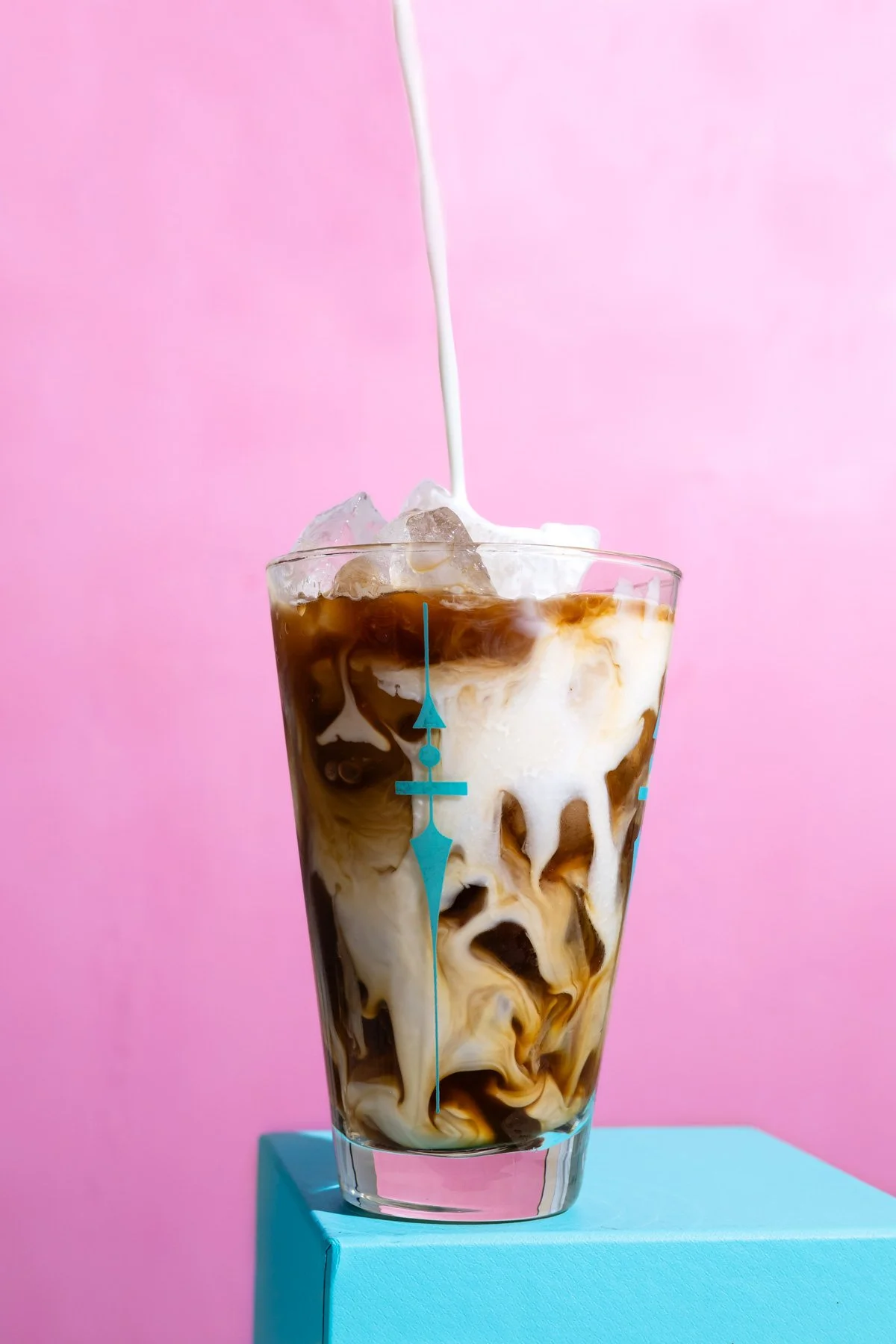 Tall glass of iced coffee with milk and chocolate swirls, ice cubes, and creamy milk being poured into it, set against a pink background.