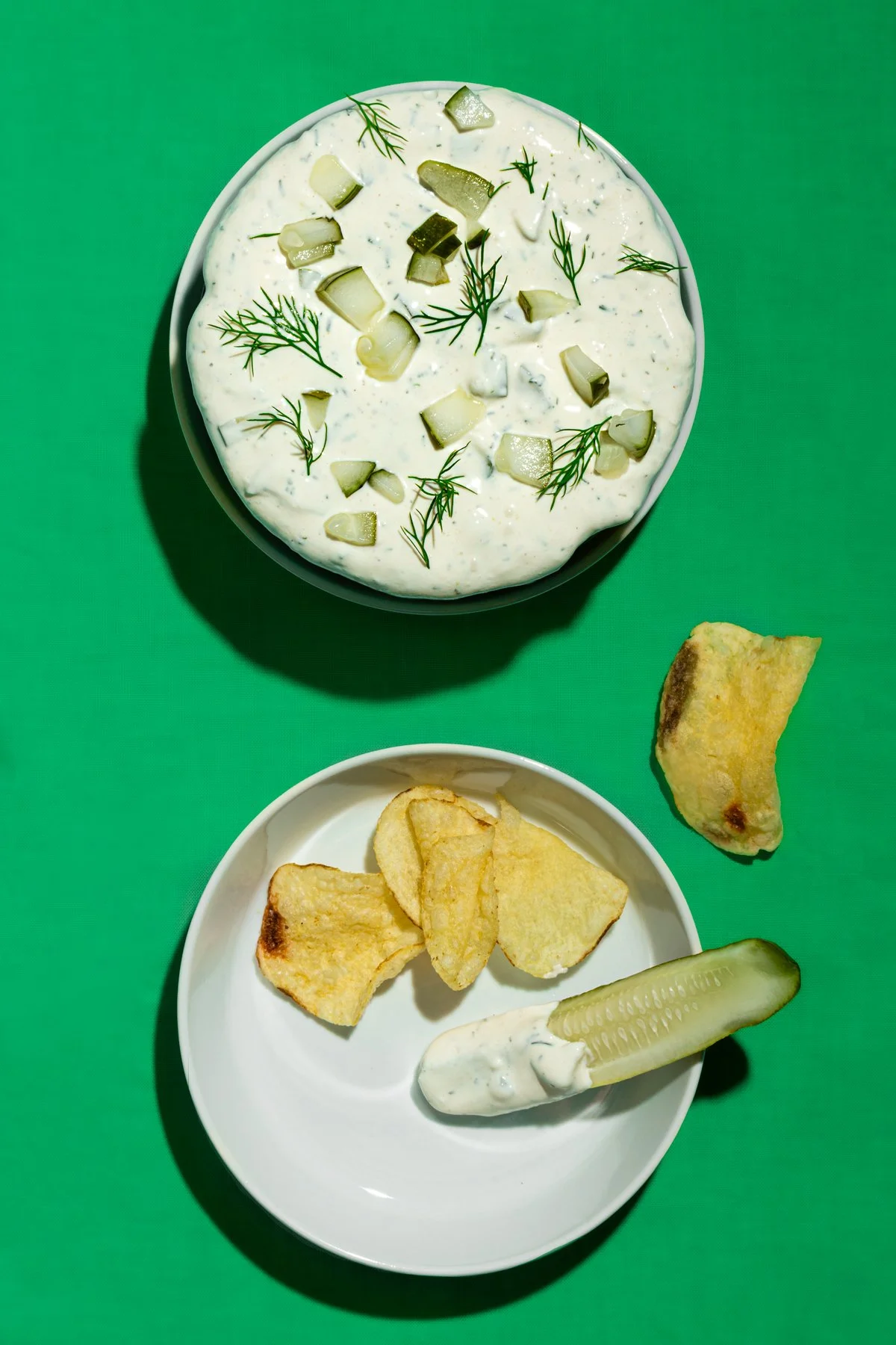 A bowl of creamy cucumber salad topped with chopped cucumbers and dill, accompanied by potato chips, a slice of cucumber, a dollop of sour cream, and a baked empanada on a green surface.