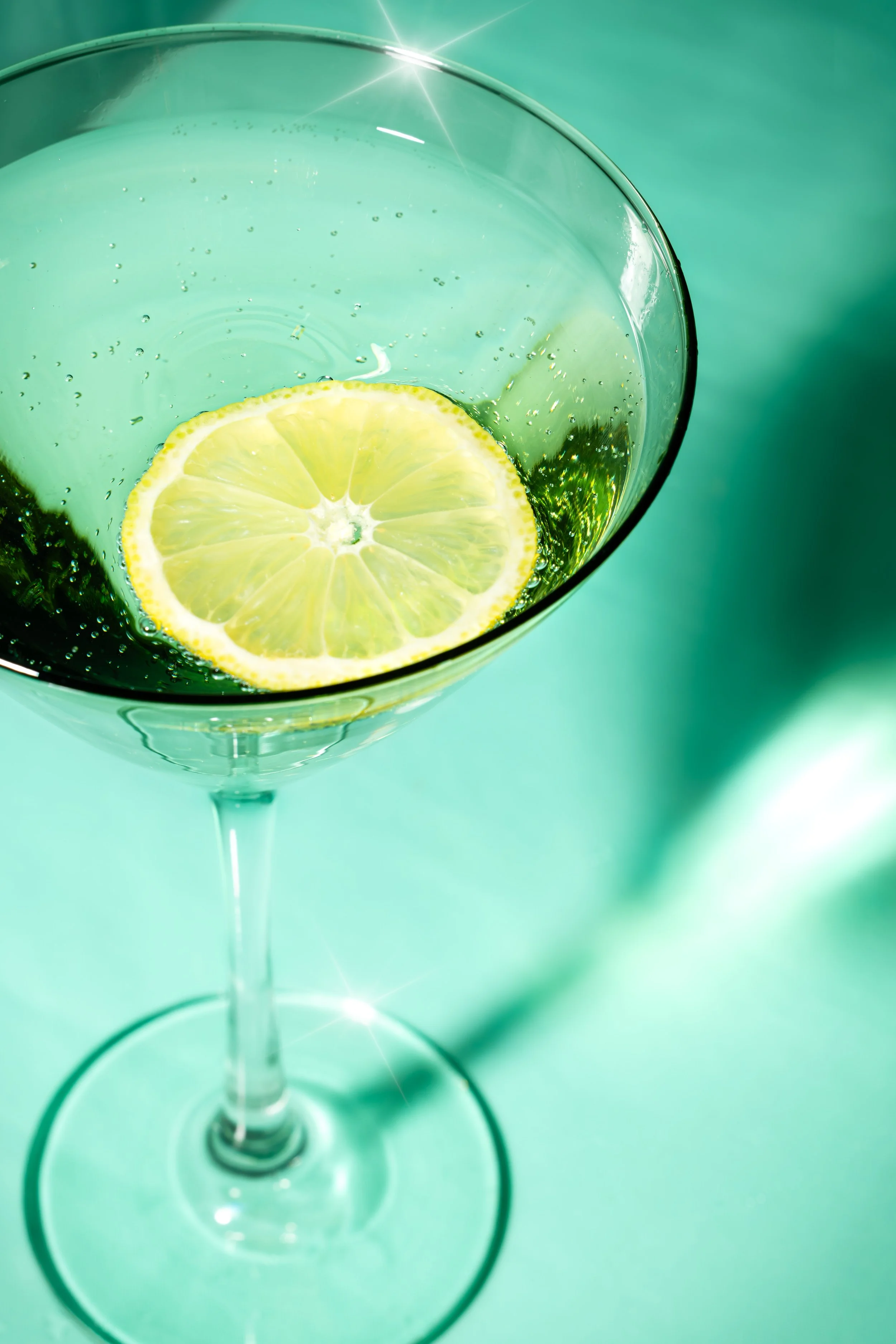 A close-up of a cocktail glass with a slice of lemon inside, filled with a bubbly, light green beverage.