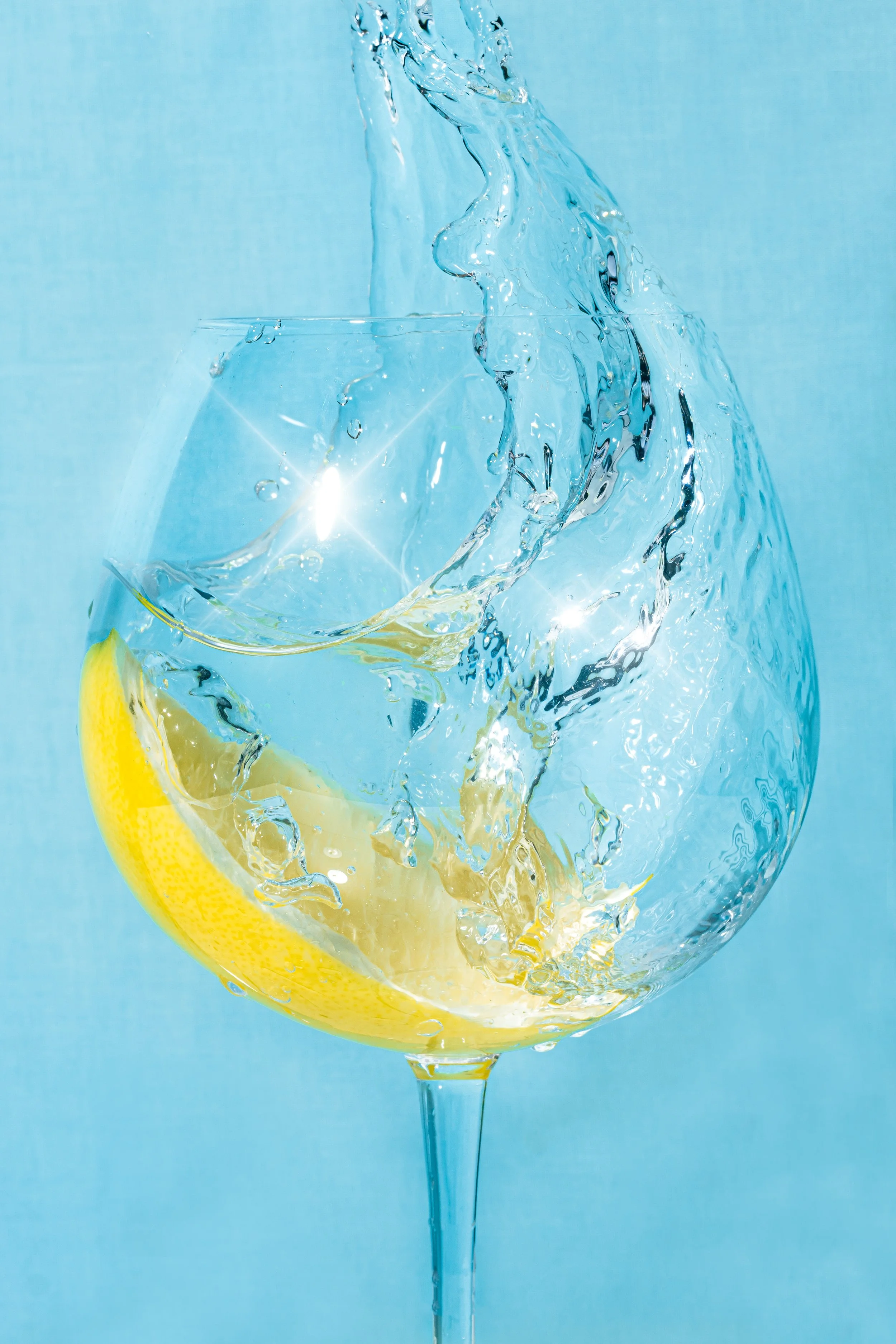 A glass of water with a lemon wedge, water splashing over the rim, and a blue background.