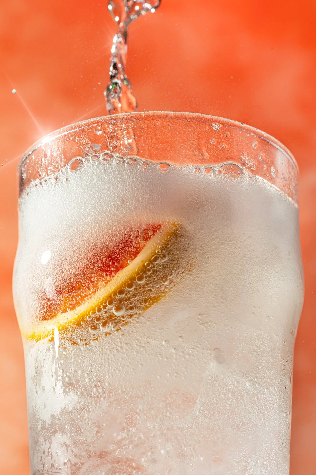 A close-up of a glass of soda with ice and a lemon wedge, with carbonated bubbles and water being poured into it.
