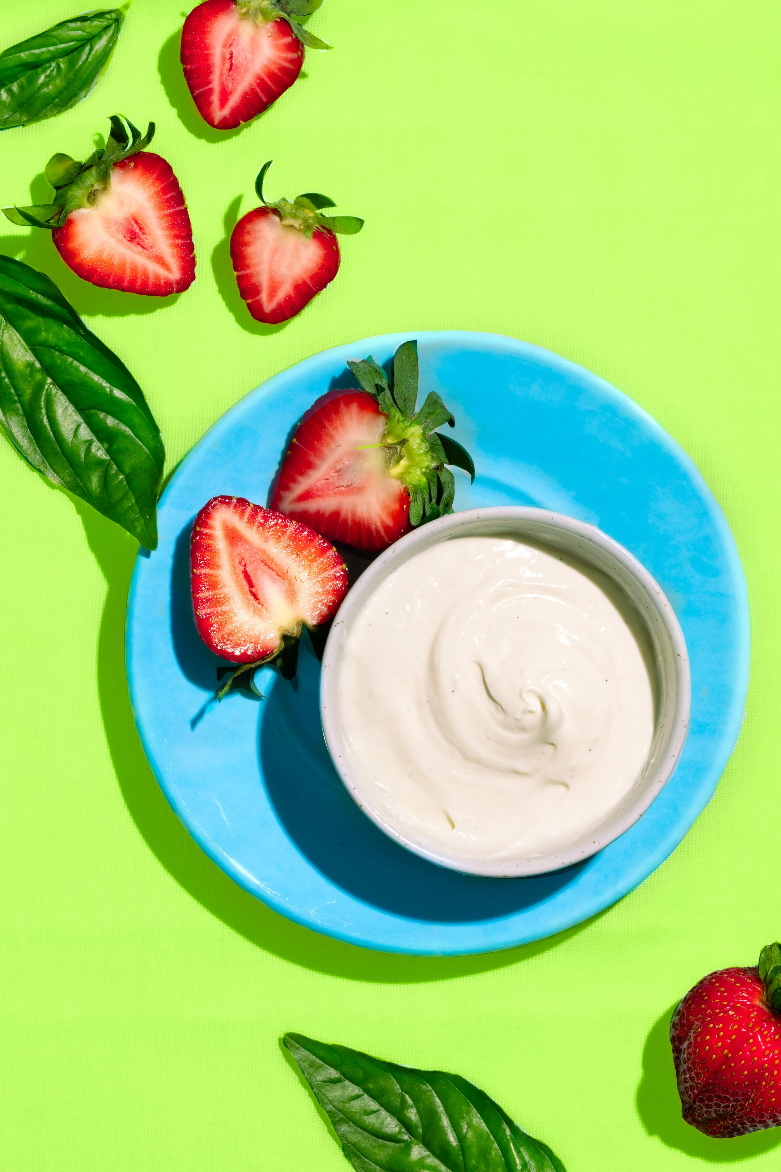 Fresh strawberries halved with green leaves and a bowl of creamy white yogurt on a bright blue plate, set against a vivid green background with scattered basil leaves.
