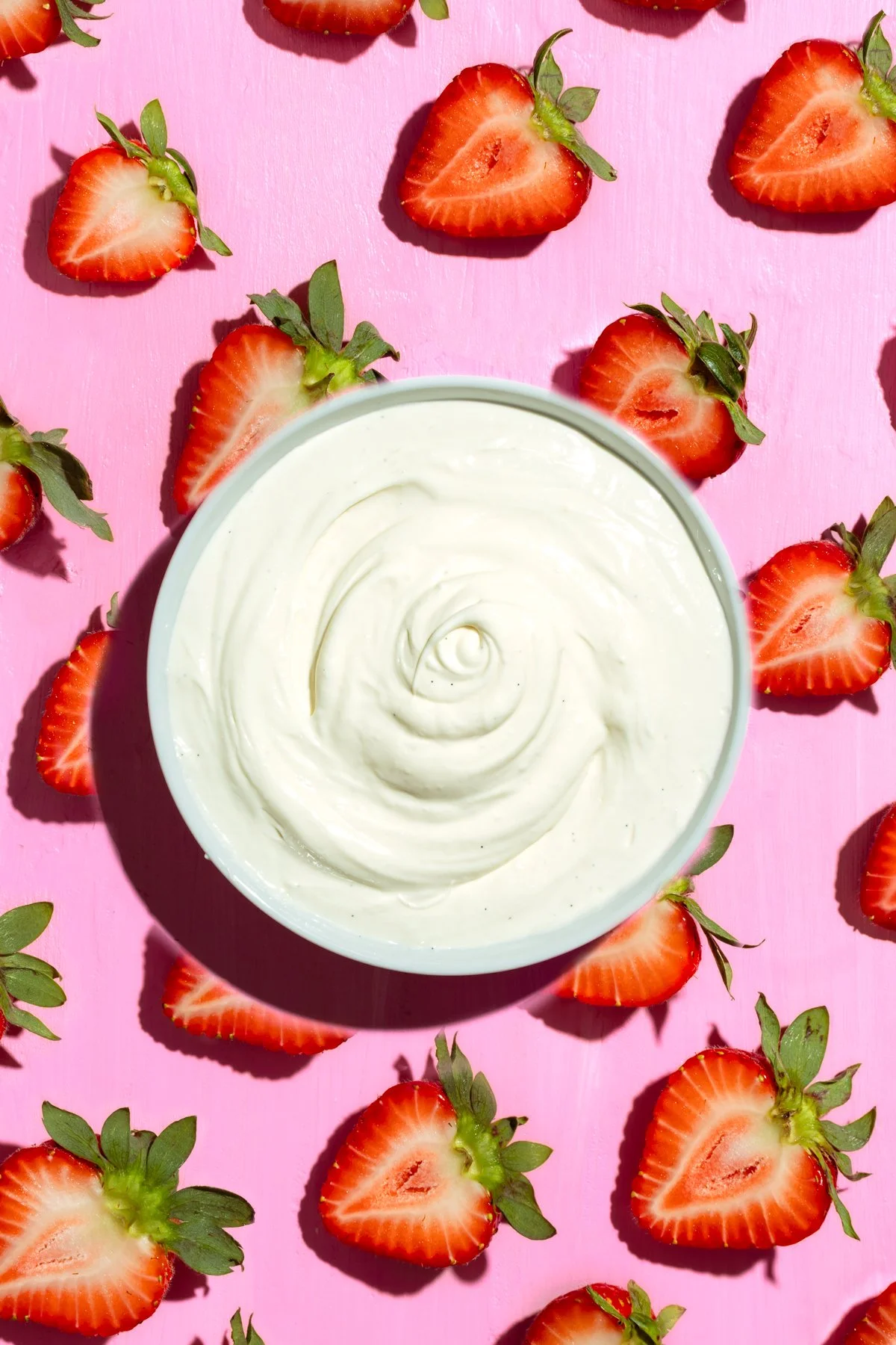 A bowl of white cream or yogurt on a pink background surrounded by sliced strawberries with green leaves.