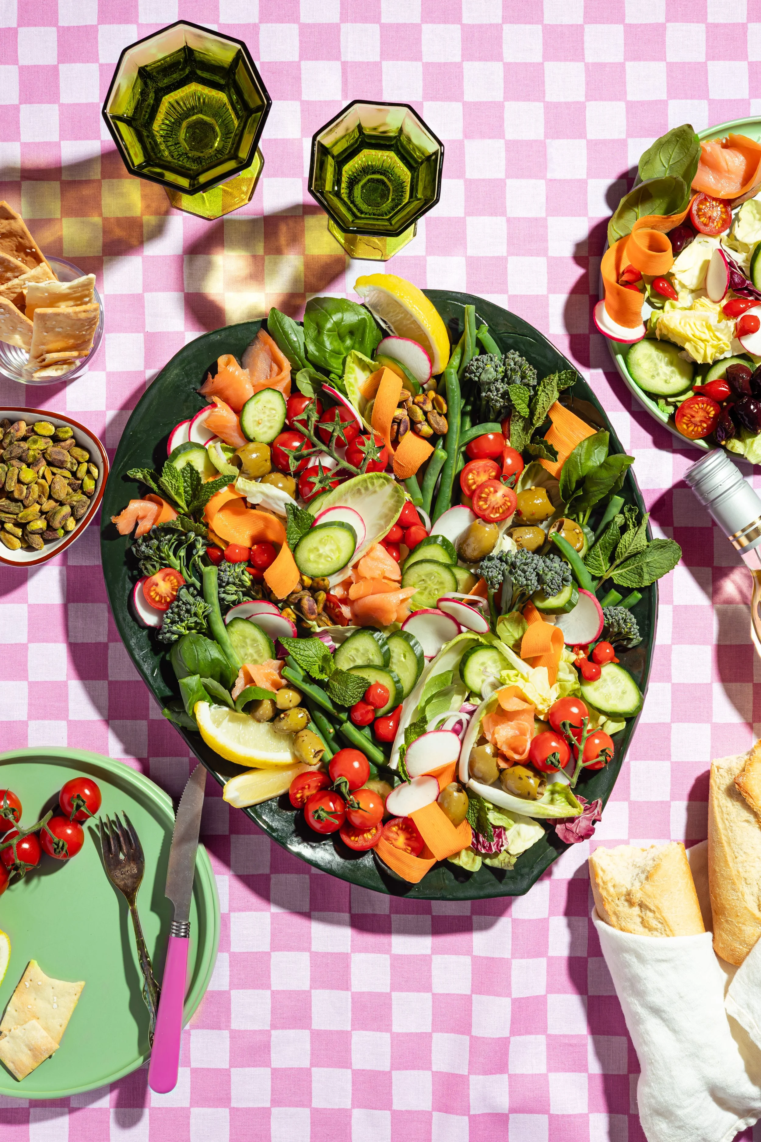 Colorful salad with cherry tomatoes, cucumber, radishes, carrots, green beans, olives, and leafy greens on a black platter, on a pink checkered tablecloth. A lemon wedge is on the salad. Two green glasses with black rims are nearby, along with a bowl