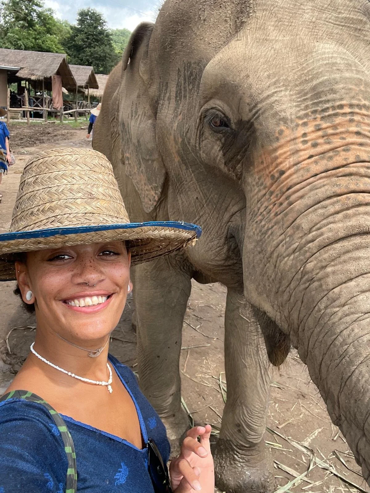 Smiling woman wearing a straw hat, white jewelry, and a blue top, taking a selfie with an elephant in the background at a zoo or wildlife park.