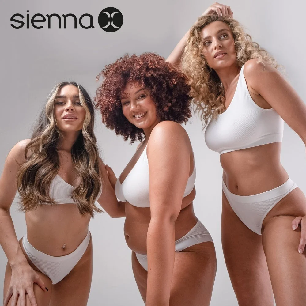 Three women posing together in white underwear, smiling, against a plain light gray background, with the 'Sienna' brand logo at the top left corner.